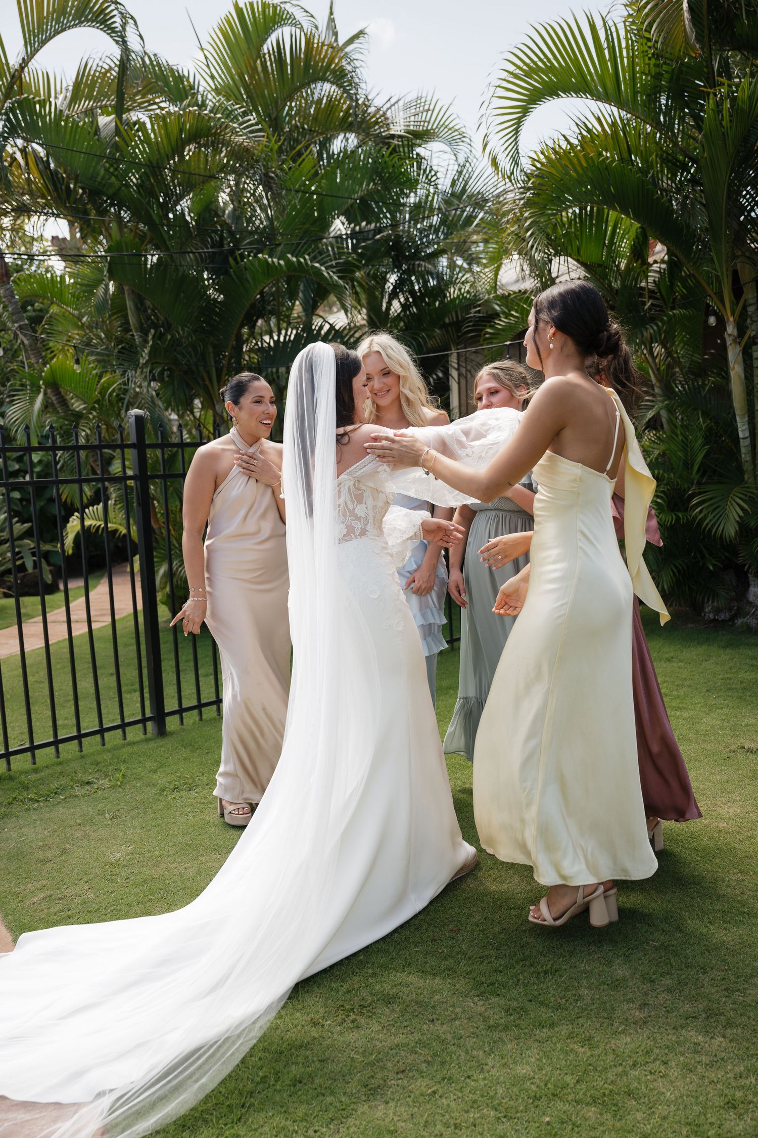 Bridesmaids in pastel dresses helping adjust a white wedding gown outdoors.