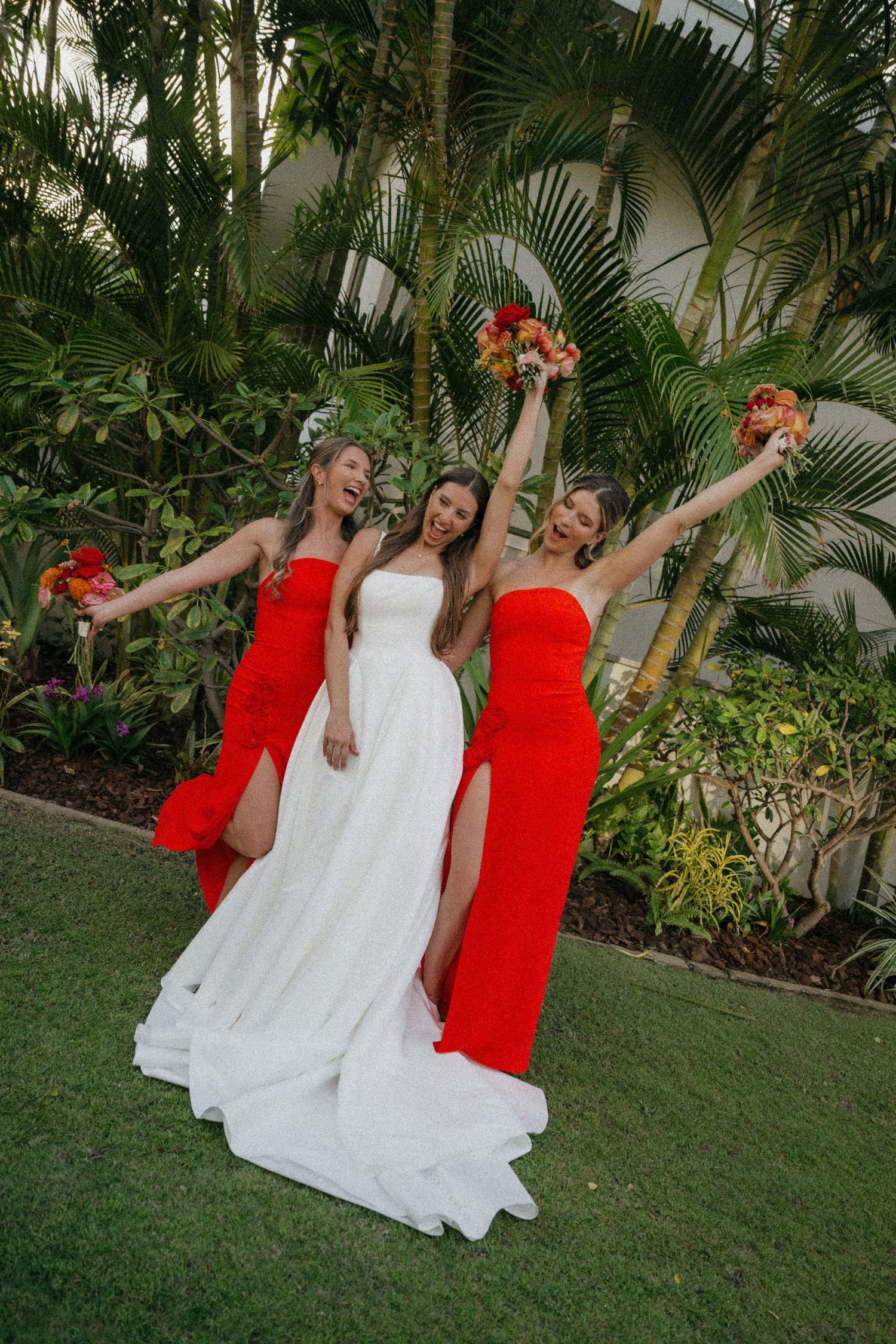 Bridal party in white dress and red dresses pose playfully on grass with tropical palm trees in background.