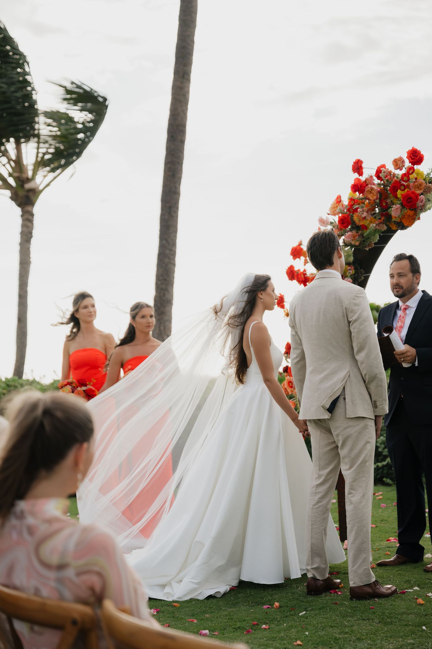 Wedding party poses for photos on a windy day with palm trees and vibrant red flowers in the background.
