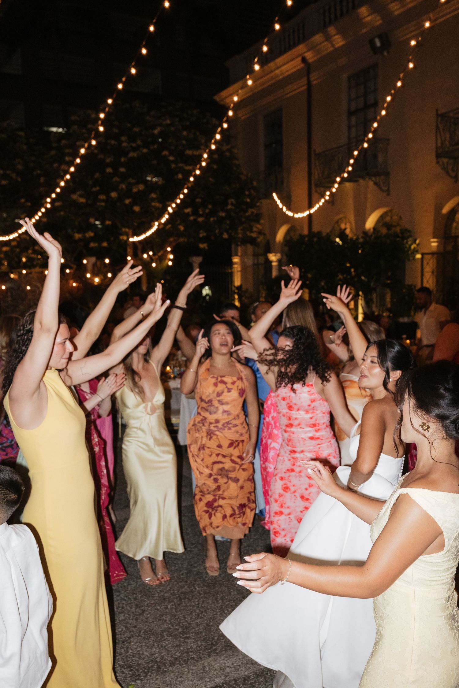 Wedding guests dancing outdoors at night under string lights, wearing colorful formal dresses with arms raised.