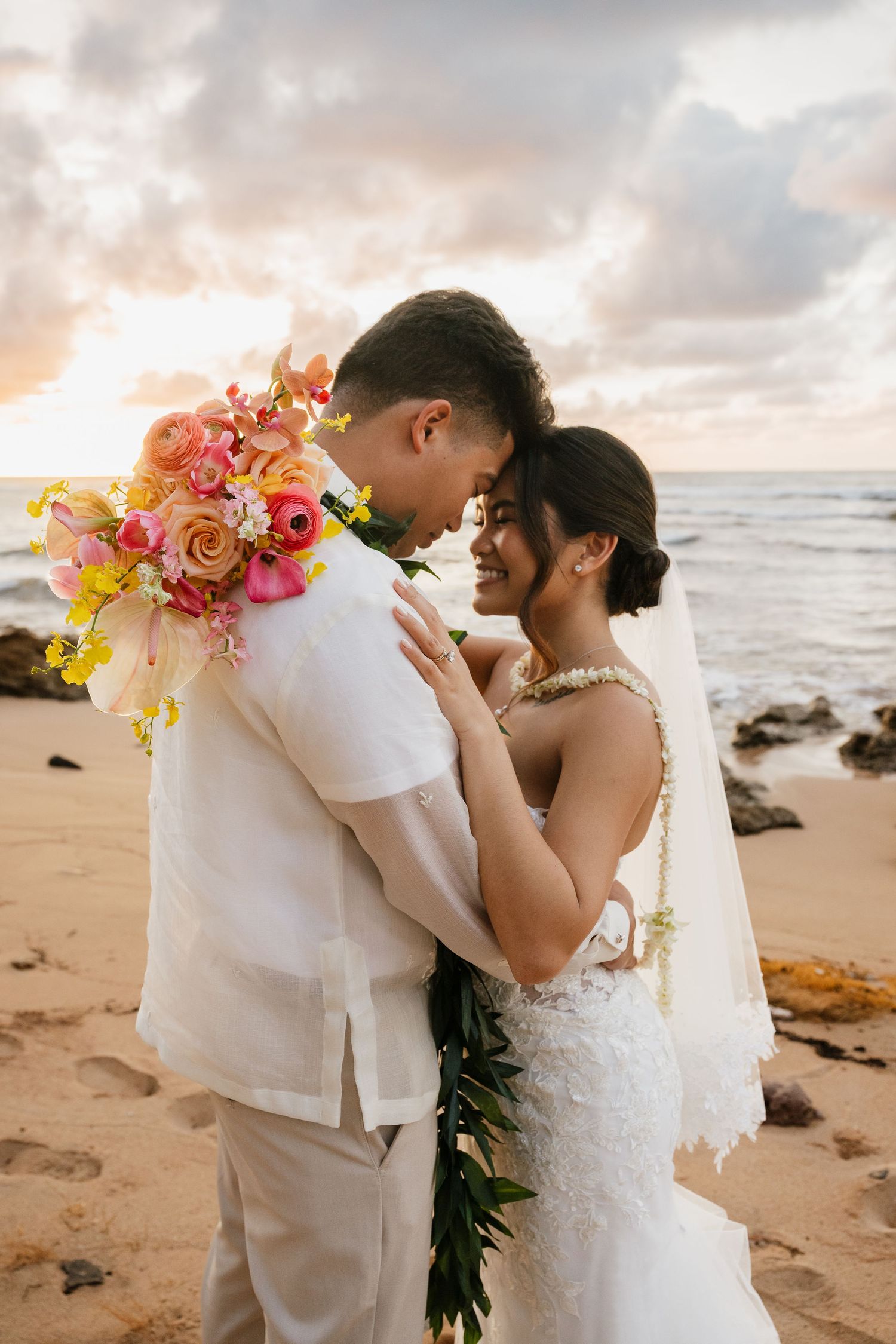 Bride and groom embrace on sandy beach at sunset holding vibrant tropical wedding bouquet.