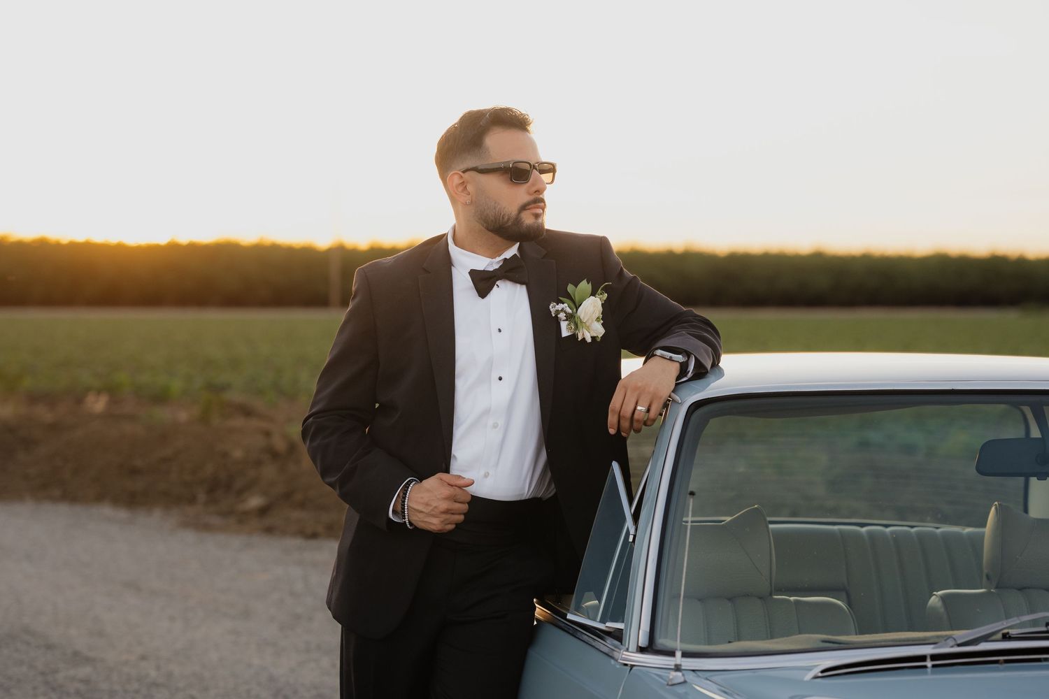 A groom stands next to a vintage car at sunset during a wedding photoshoot.