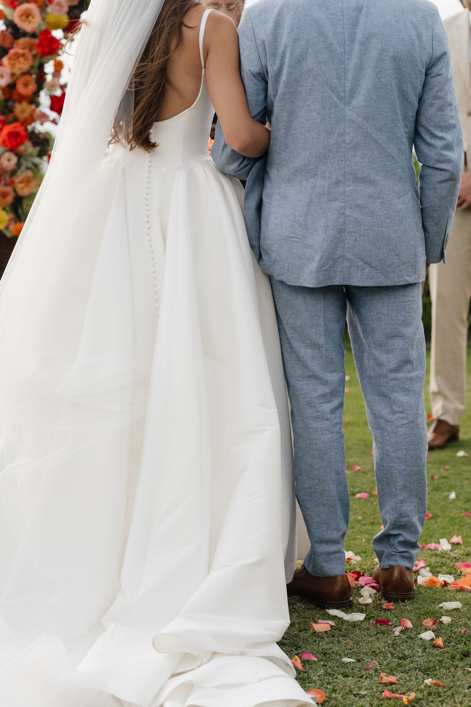 A wedding couple stands together on grass scattered with rose petals during their outdoor ceremony.