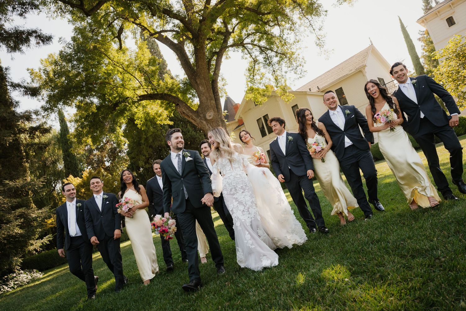 Wedding party members walk and laugh together on a grassy lawn during an outdoor celebration.