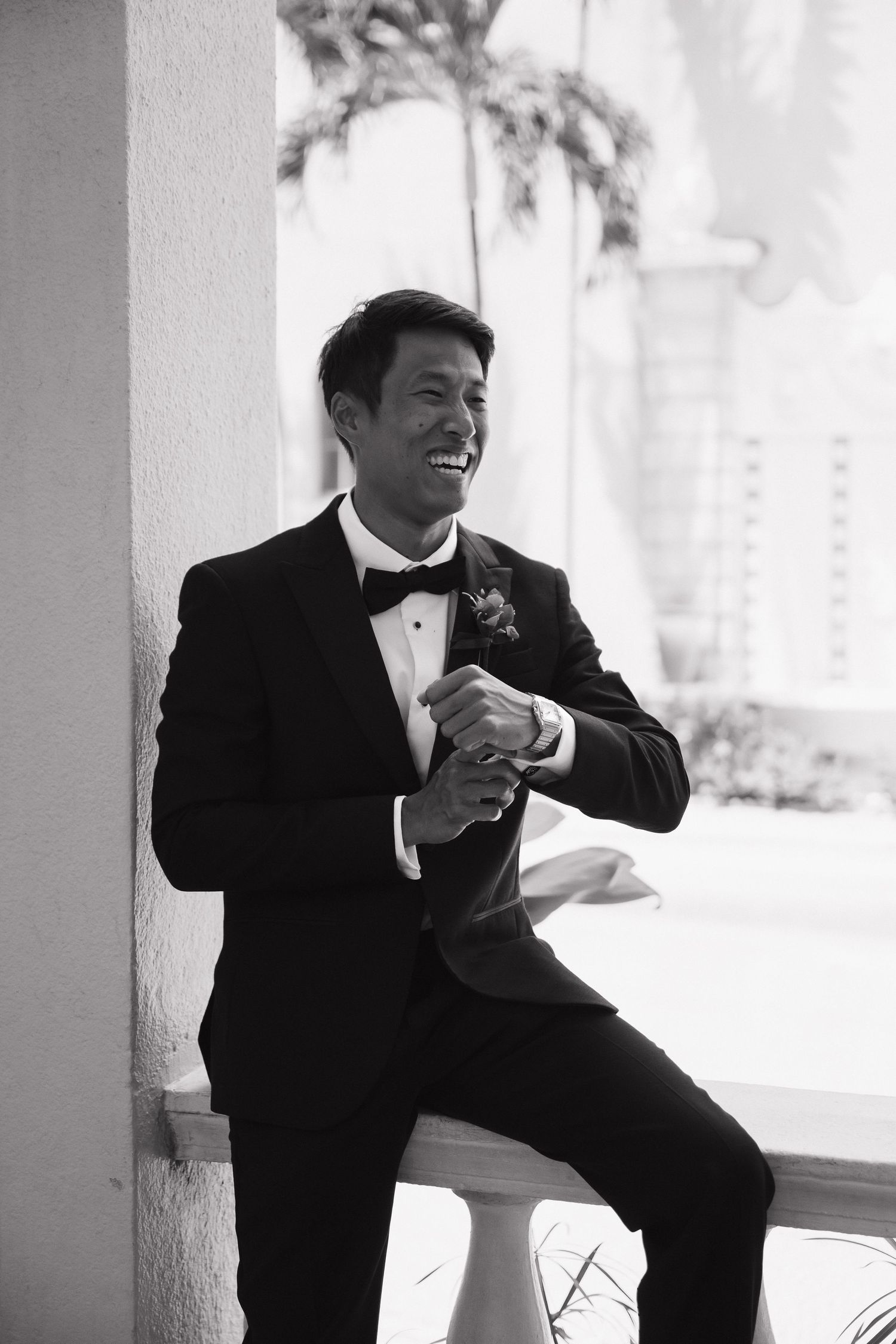 Smiling groom in black tuxedo with boutonniere leans against white column outdoors in black and white photo.