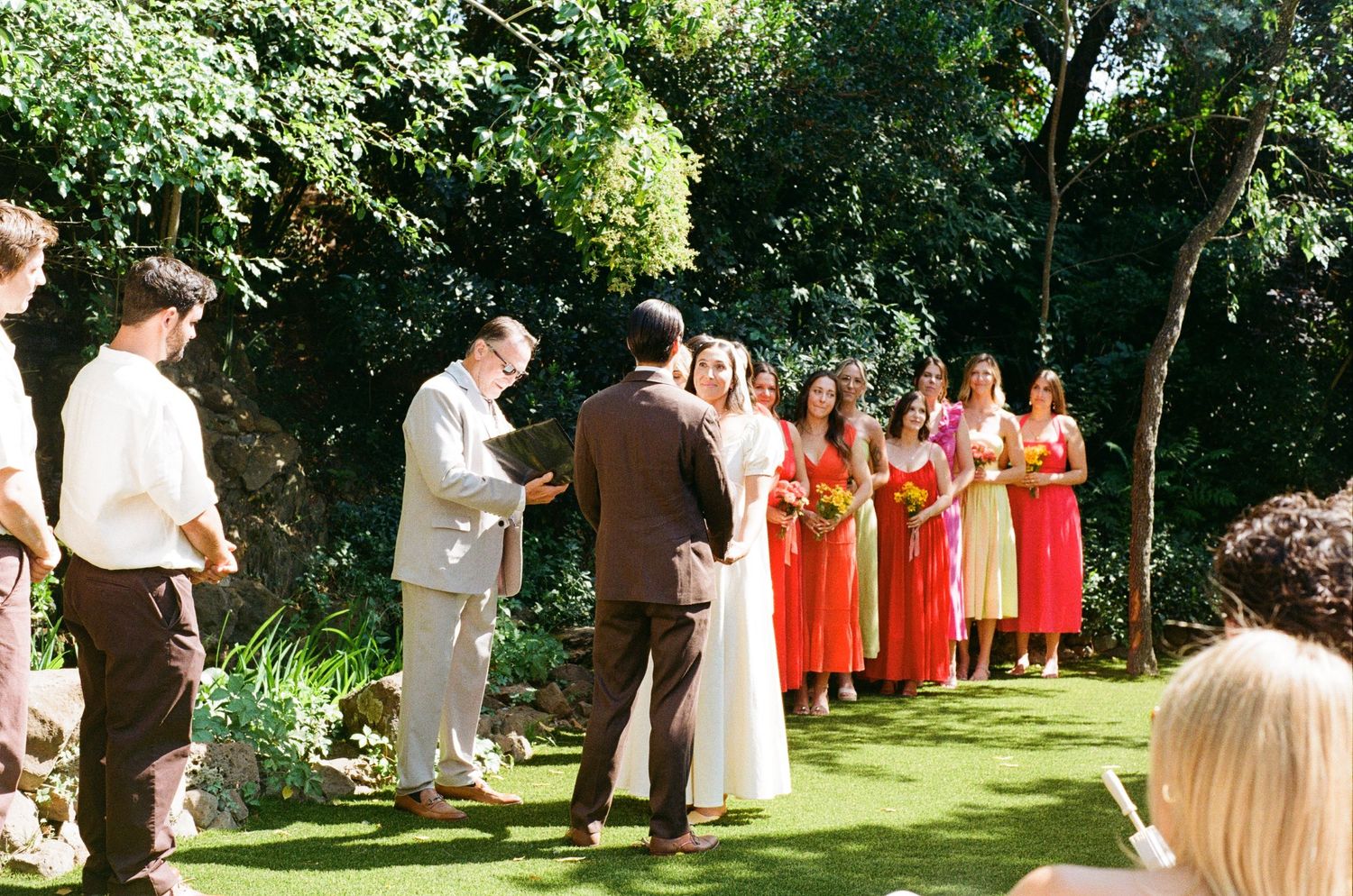 An outdoor wedding ceremony takes place in a lush garden setting with bridesmaids wearing bright coral dresses.