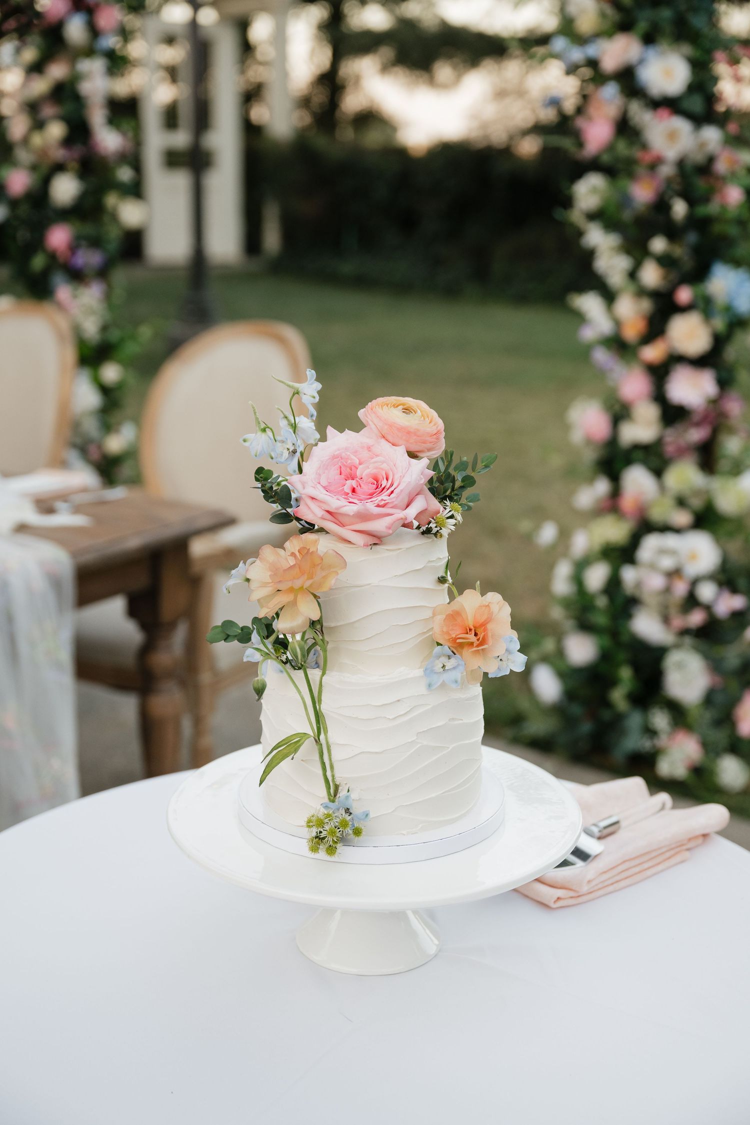 White textured wedding cake decorated with fresh pink and peach flowers on a white cake stand.