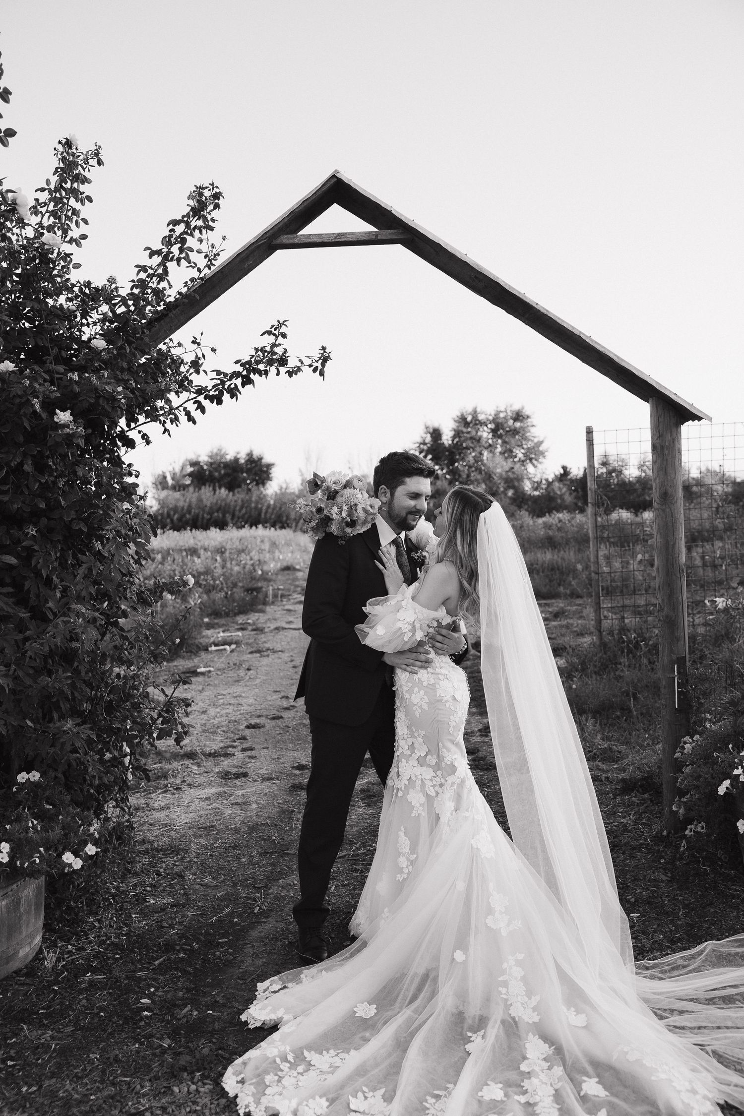A romantic wedding moment captured in black and white under a wooden arch in a garden setting.