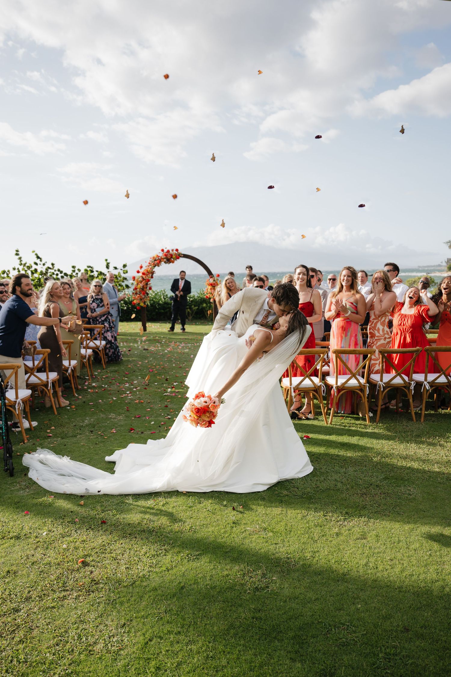 Rose petals float through the air at an outdoor wedding ceremony as a bride's veil flows dramatically in the wind.