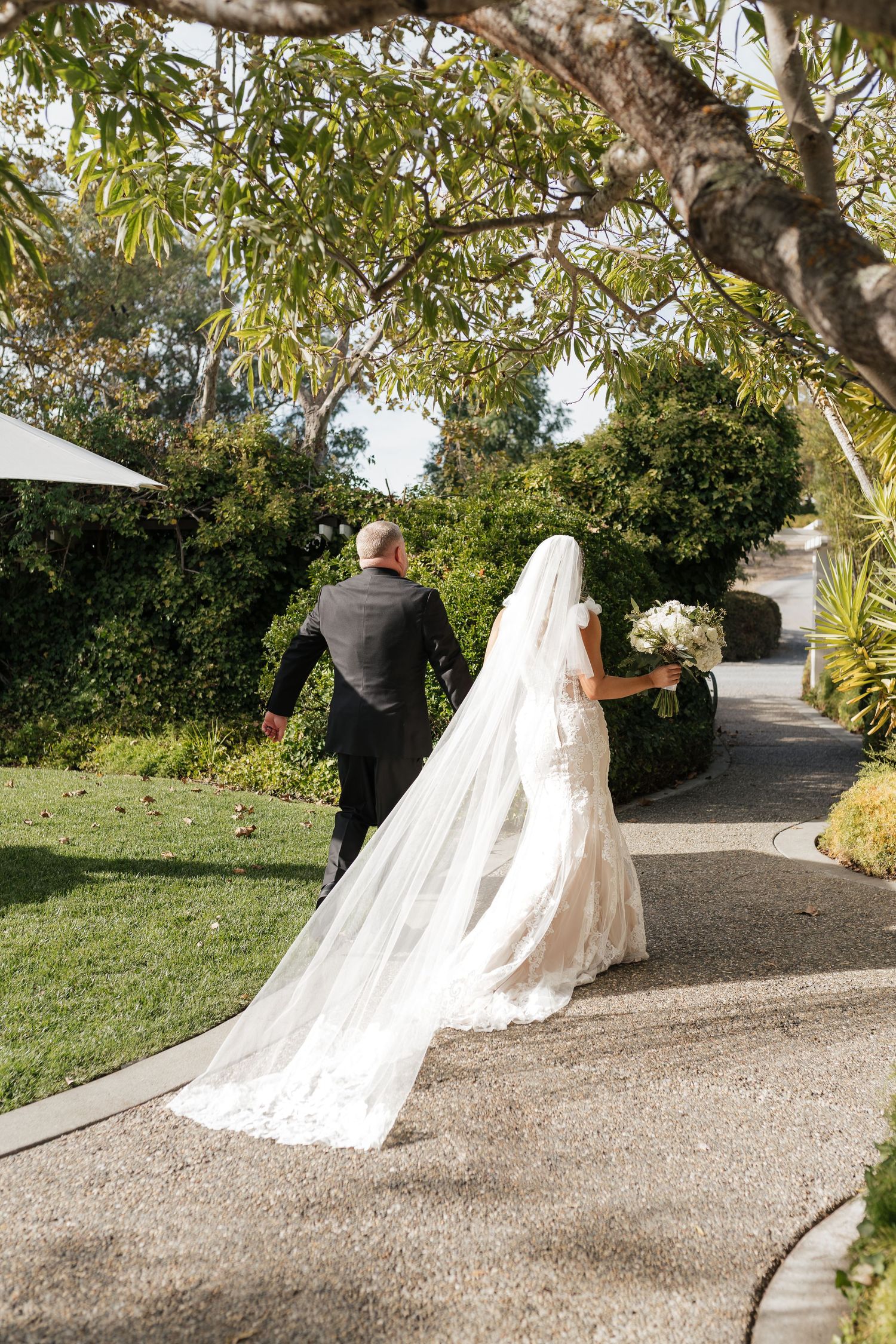 Couple walking down garden path with flowing white wedding dress and veil trailing behind.