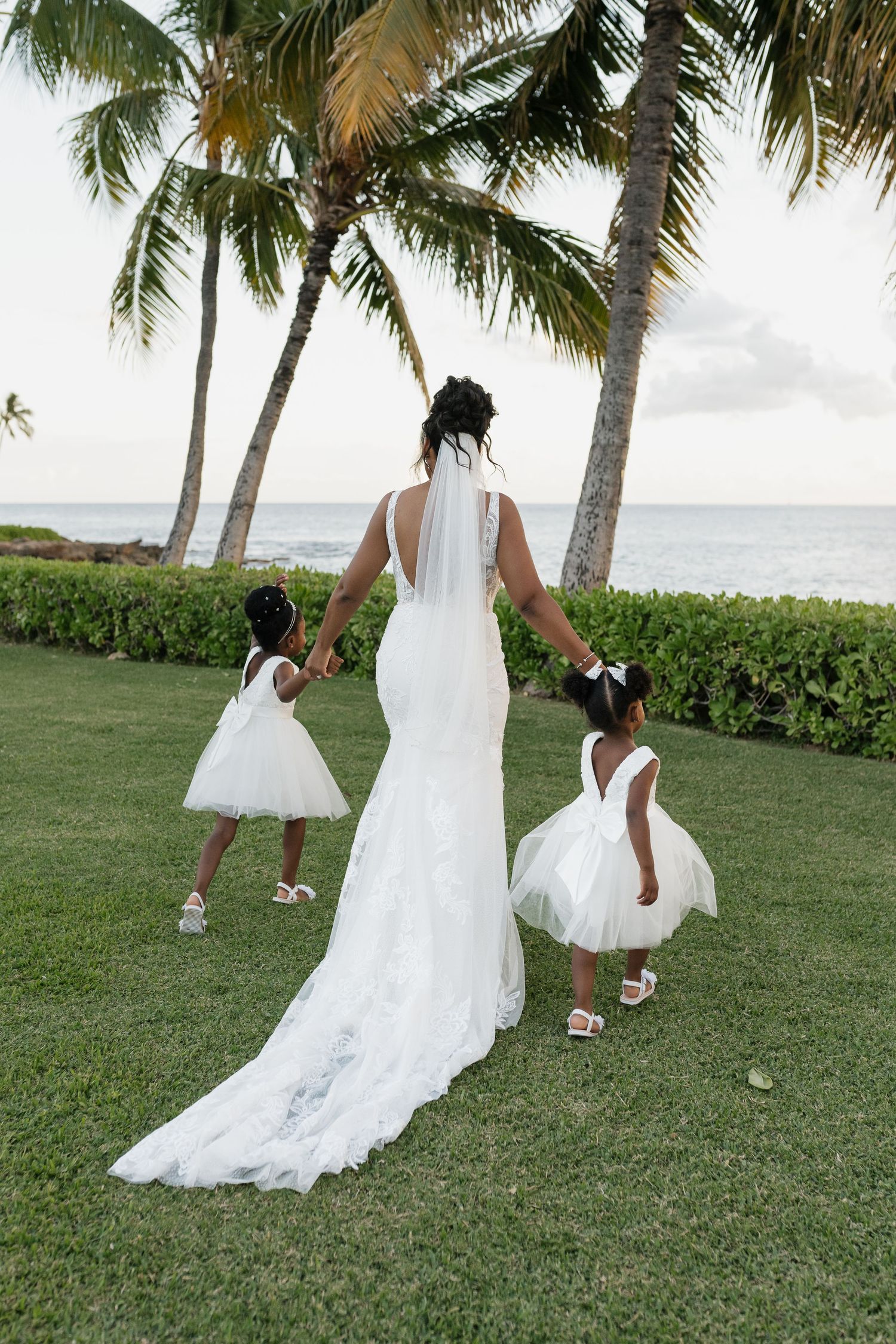 Wedding dress trains flow across green lawn with palm trees and ocean view.