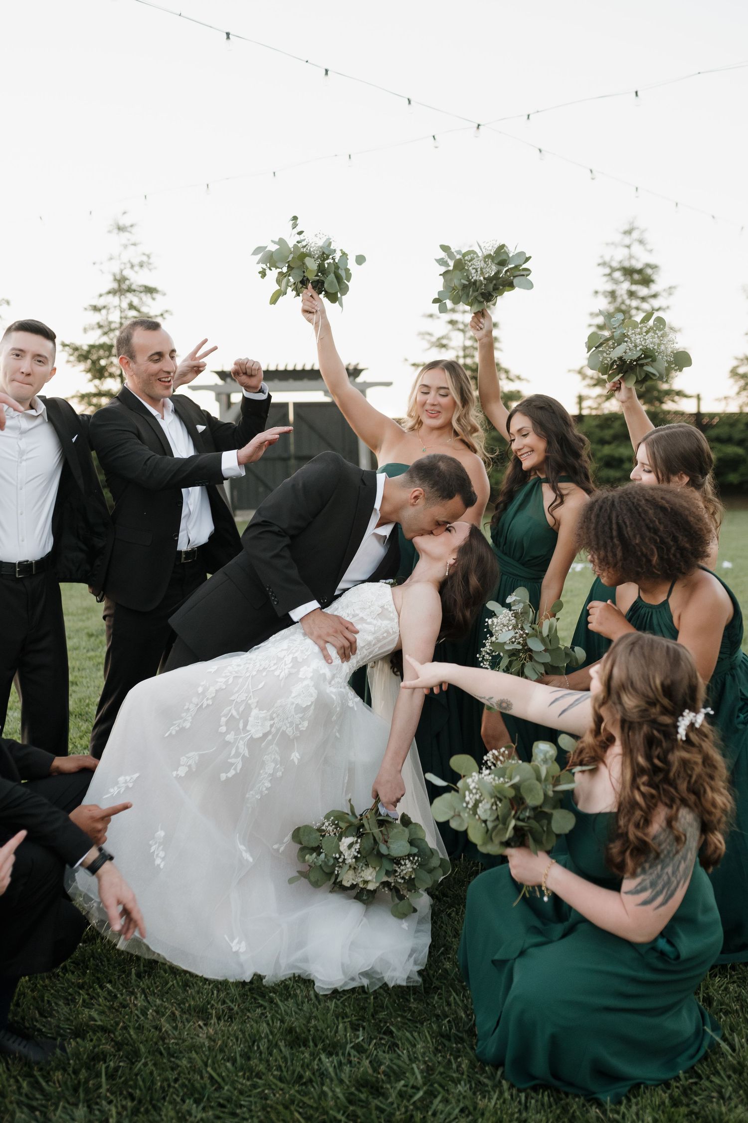 Wedding party relaxes on grass in emerald green dresses and formal attire during outdoor celebration.