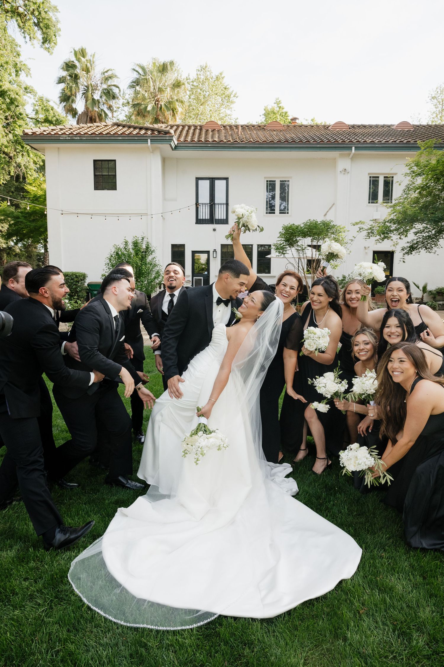 Bride and groom celebrate with wedding party in black attire on green lawn outside white Spanish-style venue.
