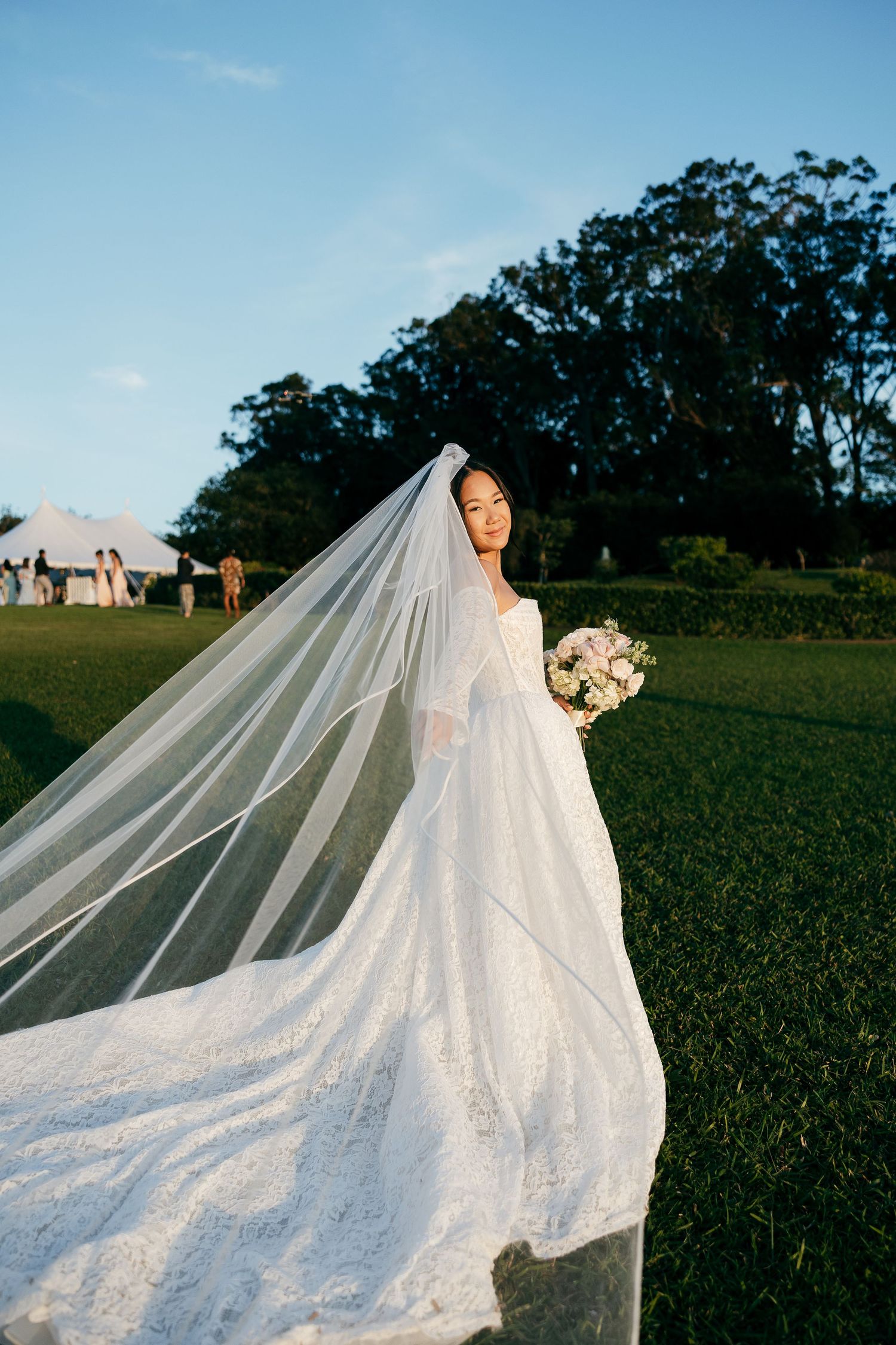 Bride in lace gown with flowing cathedral veil billowing in breeze holds bouquet on green lawn.