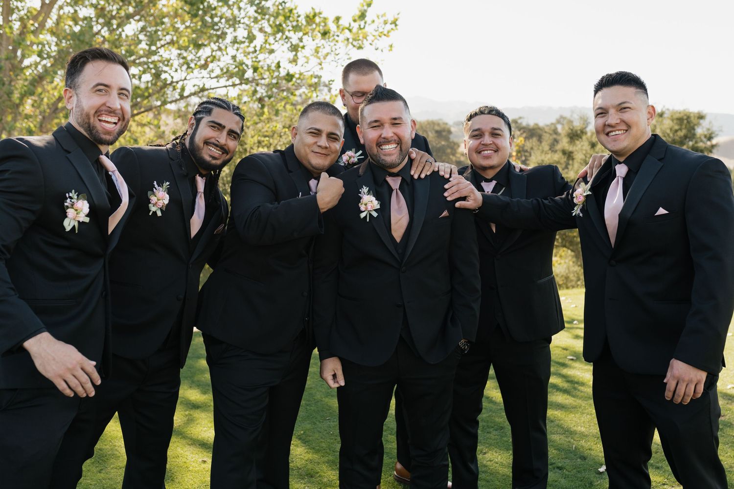 Groomsmen in black suits pose together outdoors with joyful expressions.