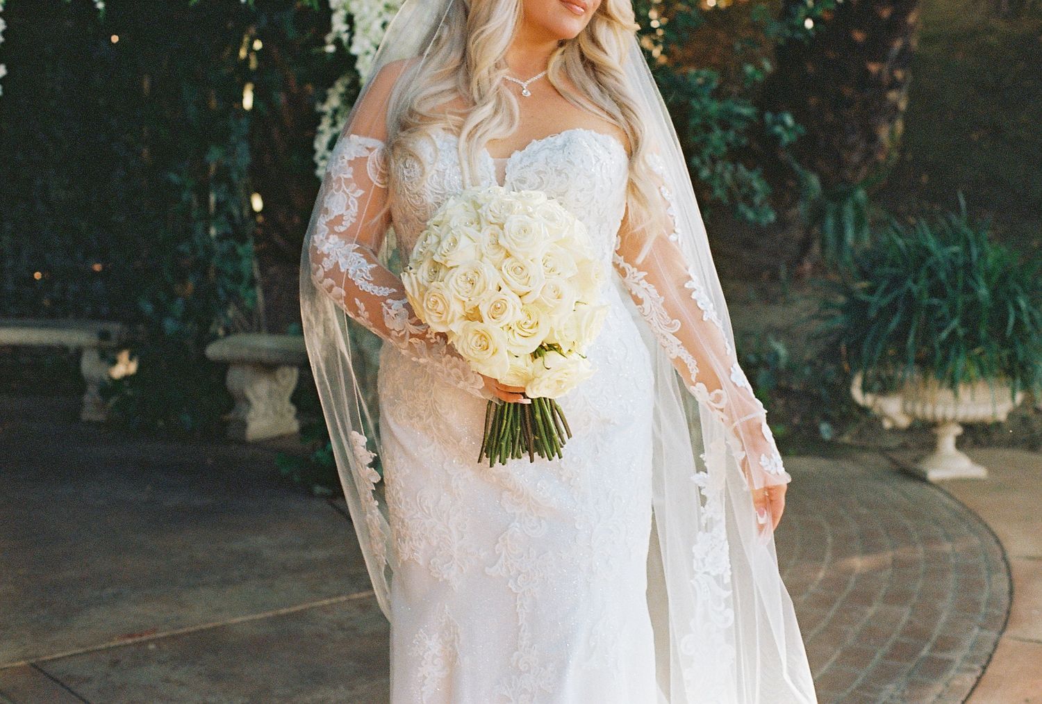 Elegant bride in lace wedding dress holding white bouquet at outdoor ceremony.