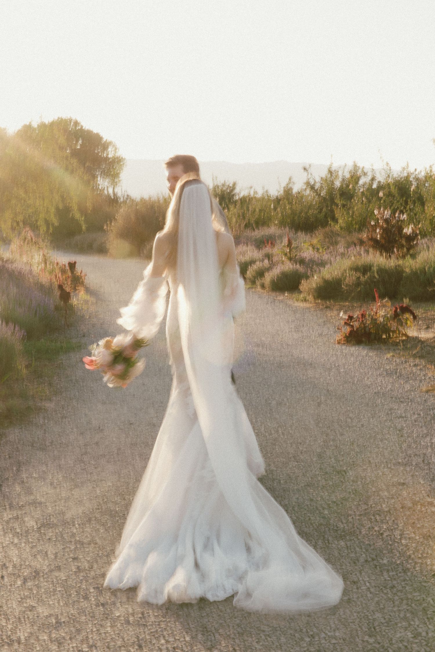 A bride in a flowing white wedding gown twirls on a path at golden hour, creating a dreamy ethereal effect.