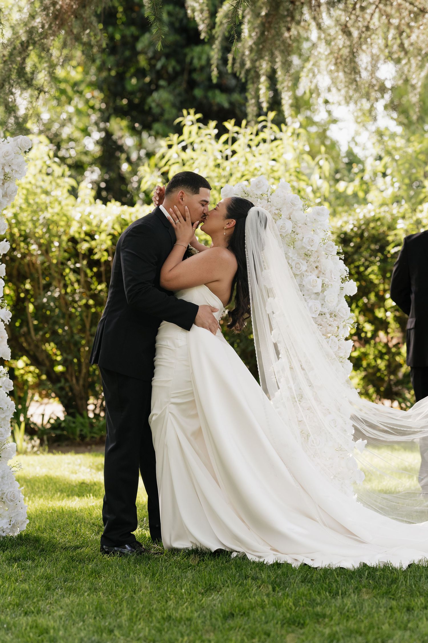Bride and groom share first kiss at outdoor wedding ceremony, bride in white gown with floral veil, groom in black suit.
