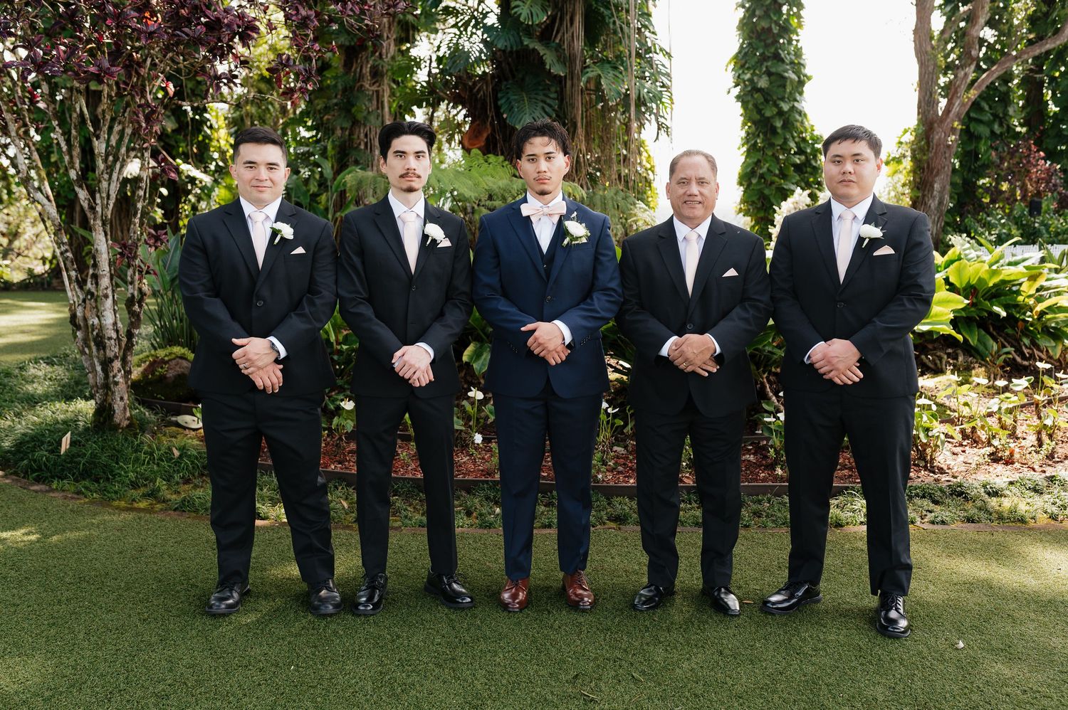 Five groomsmen in black suits pose outdoors in a lush garden setting for a formal wedding photo.