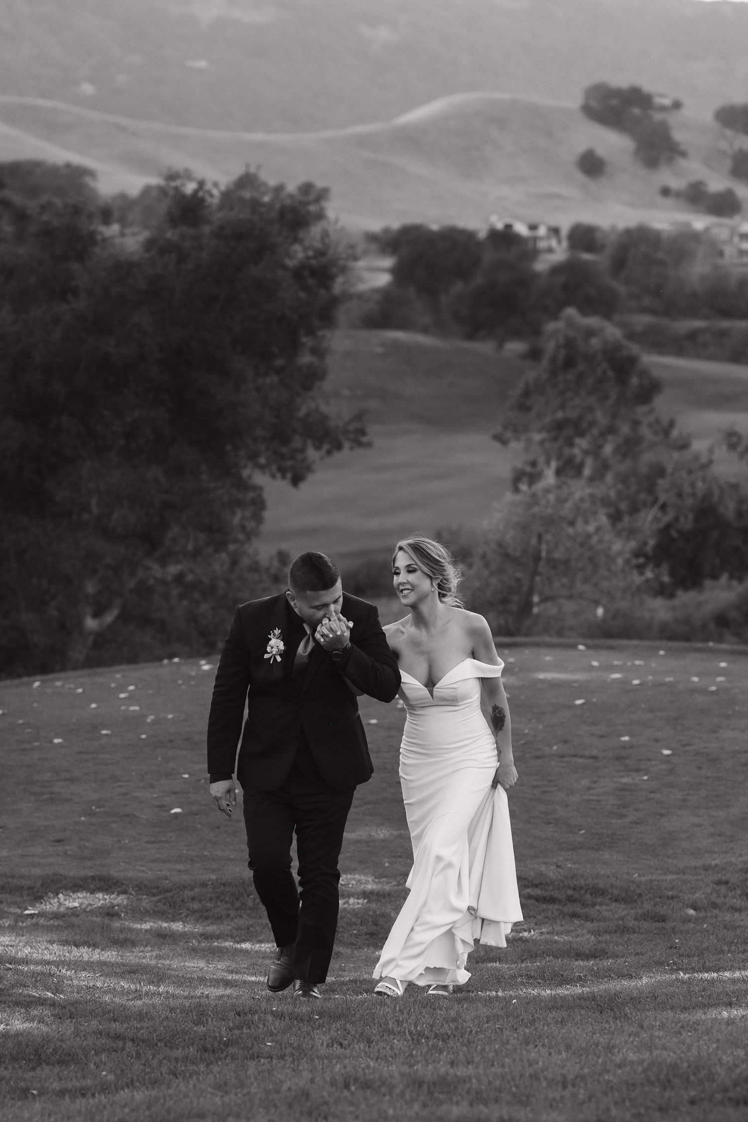 Newlyweds walk hand in hand across a scenic hilltop meadow in black and white.