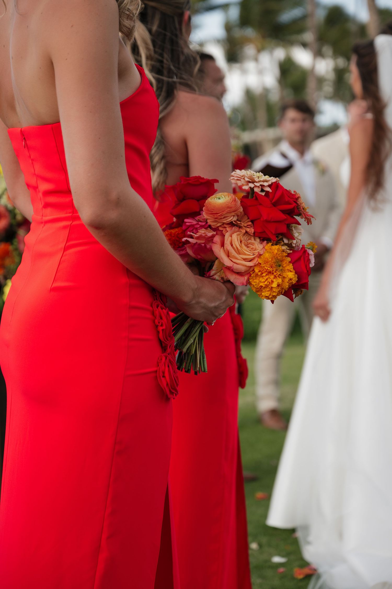 Bridesmaids in vibrant red dresses hold orange and red bouquets during an outdoor wedding ceremony.