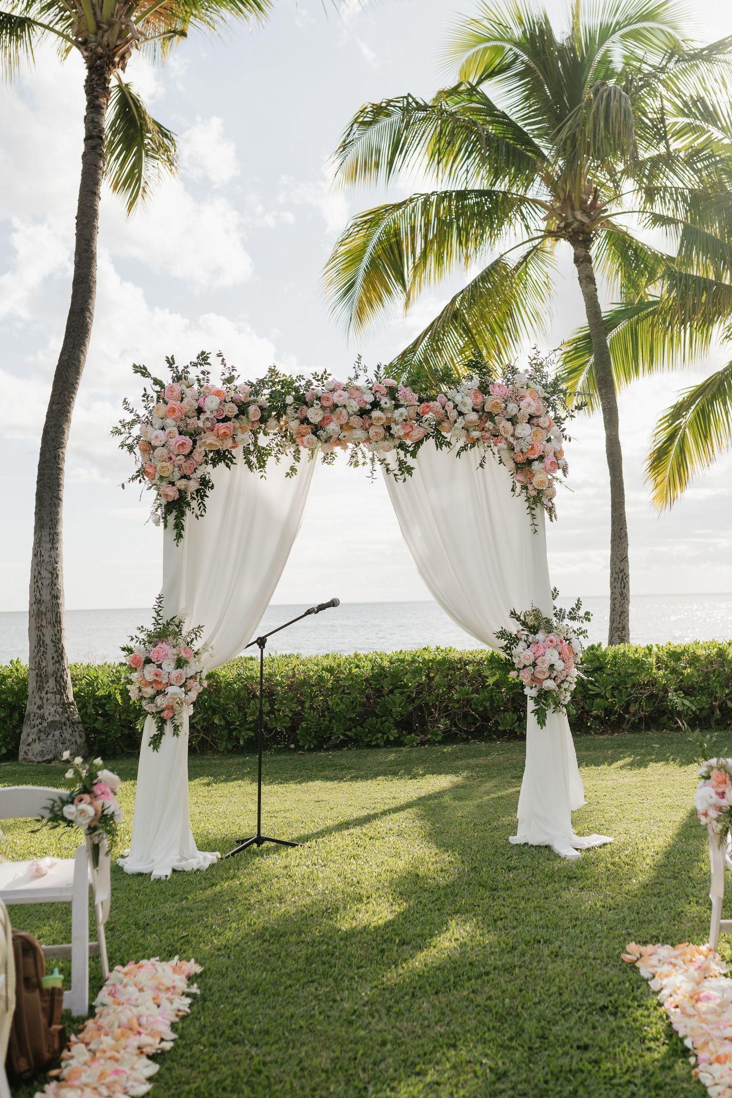 Elegant outdoor wedding ceremony setup with white drapes and pink floral arrangements under palm trees.