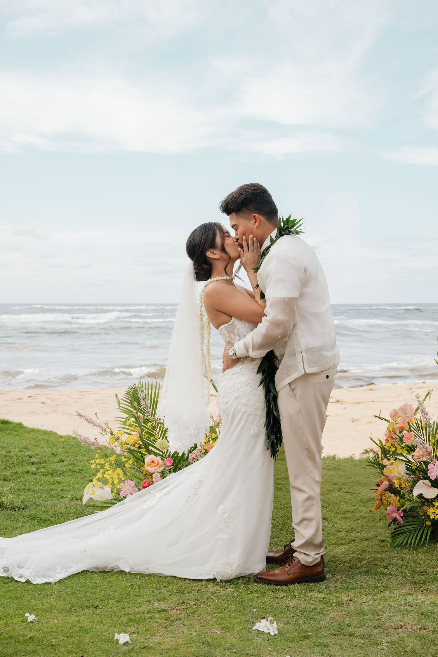 Bride and groom share first kiss at tropical beach wedding ceremony with colorful floral arrangements and ocean backdrop.