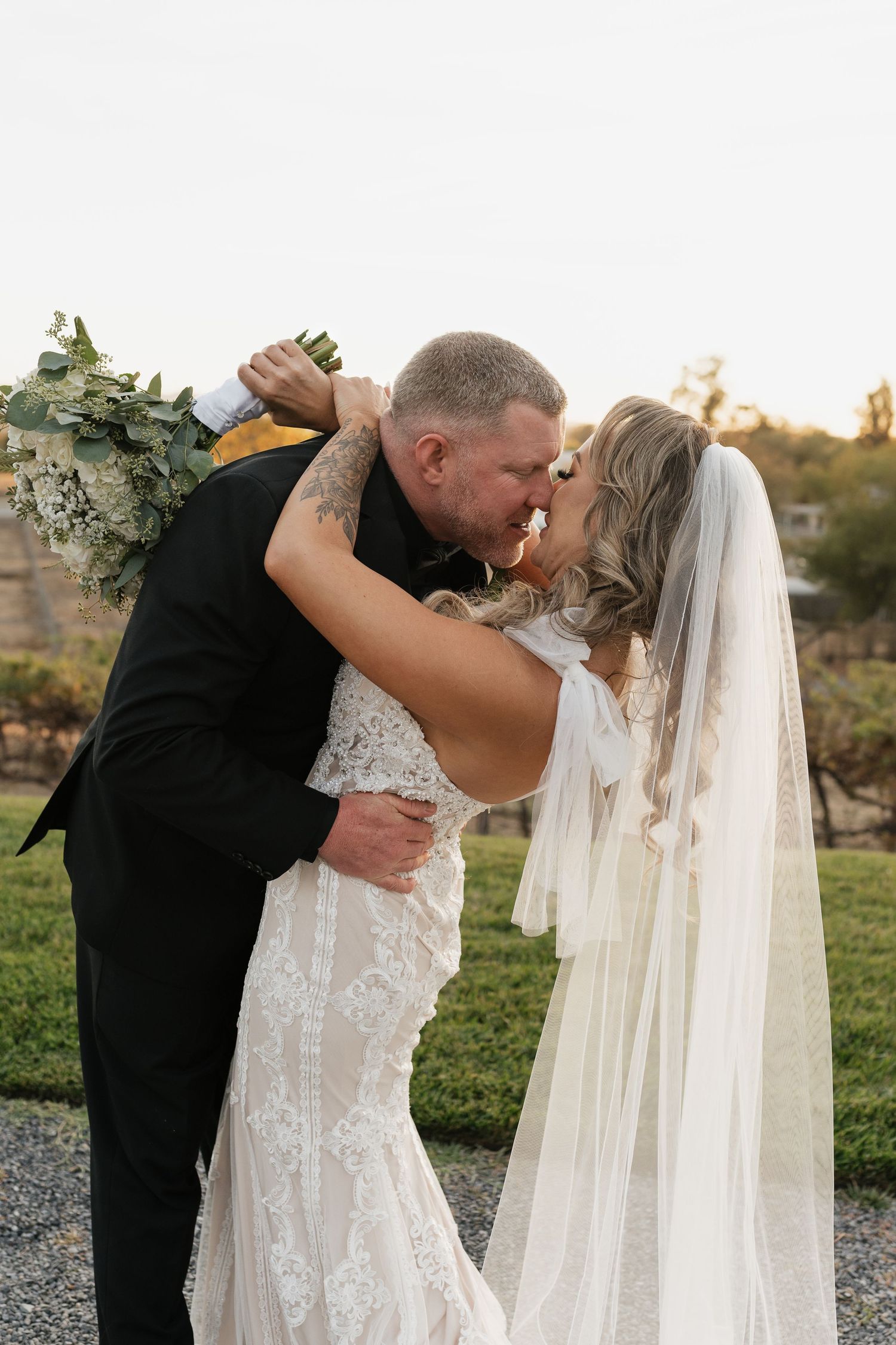 Newlyweds share an intimate kiss outdoors with the bride wearing a detailed lace gown and flowing veil.