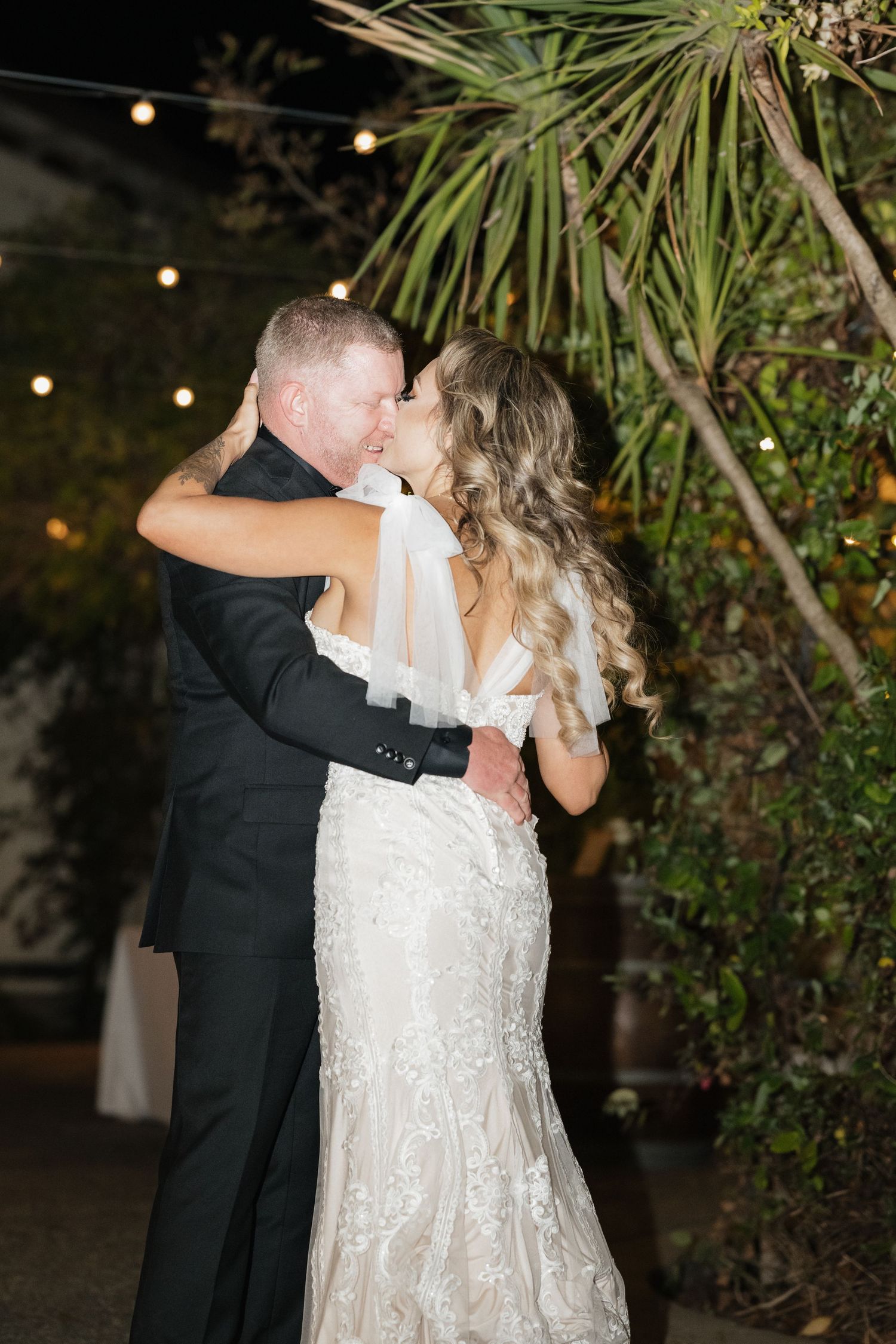 A romantic wedding kiss captured under twinkling string lights and palm trees.