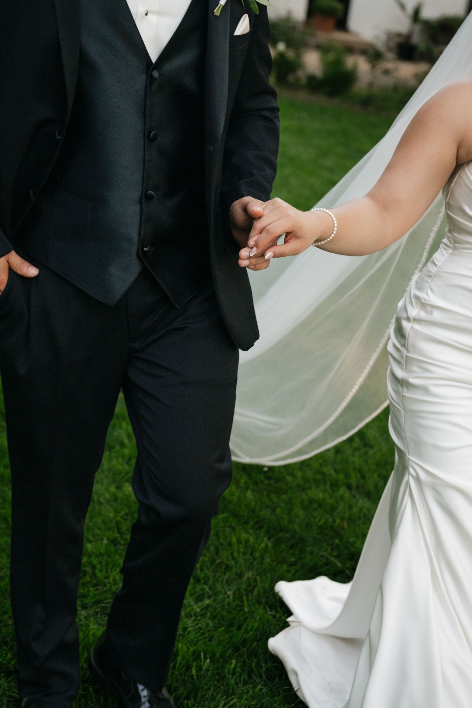 Bride and groom holding hands outdoors, wearing white wedding gown with veil and black tuxedo on green grass.