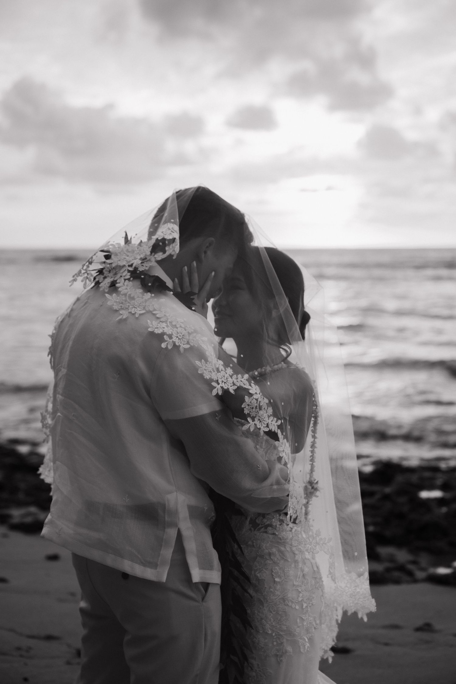 Black and white photo of bride and groom sharing intimate moment under veil on rocky beach at sunset.