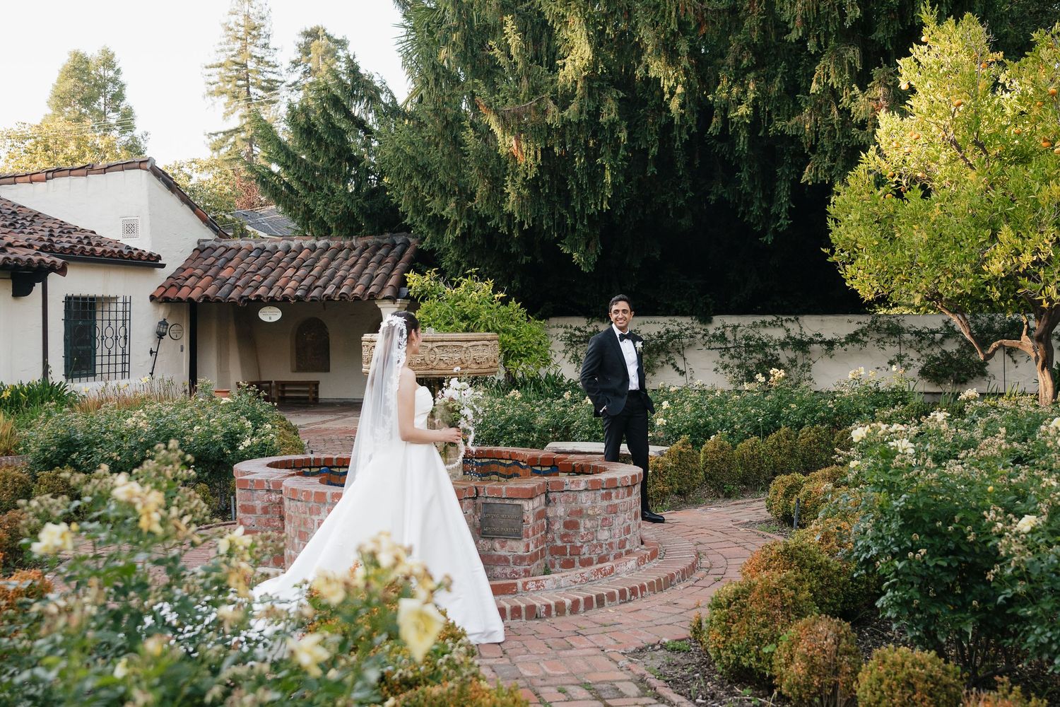 Bride and groom in formal attire stand in a lush garden courtyard near a brick fountain at a Spanish-style venue.