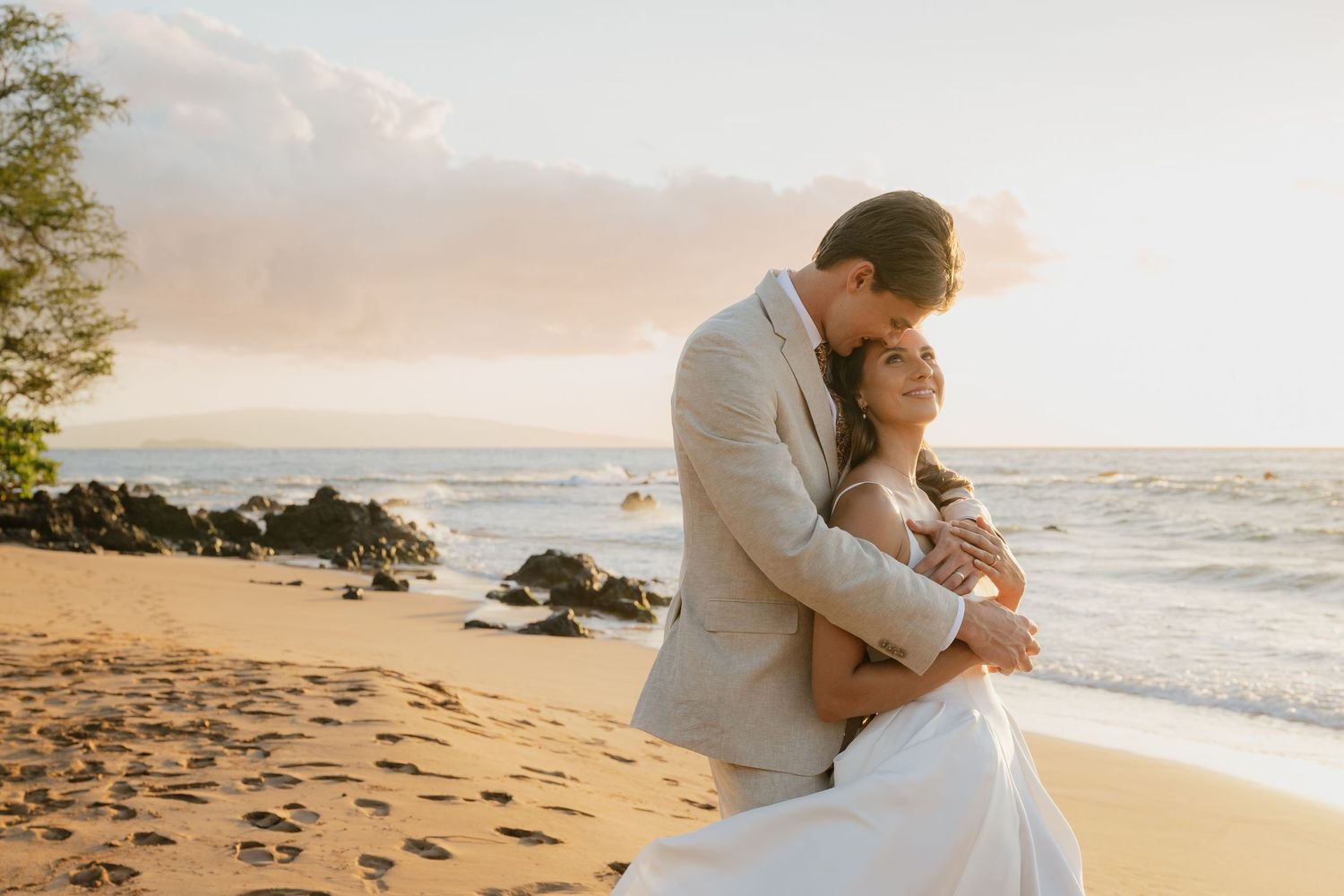 Romantic couple embracing on sandy beach at sunset with ocean waves and golden light in background.