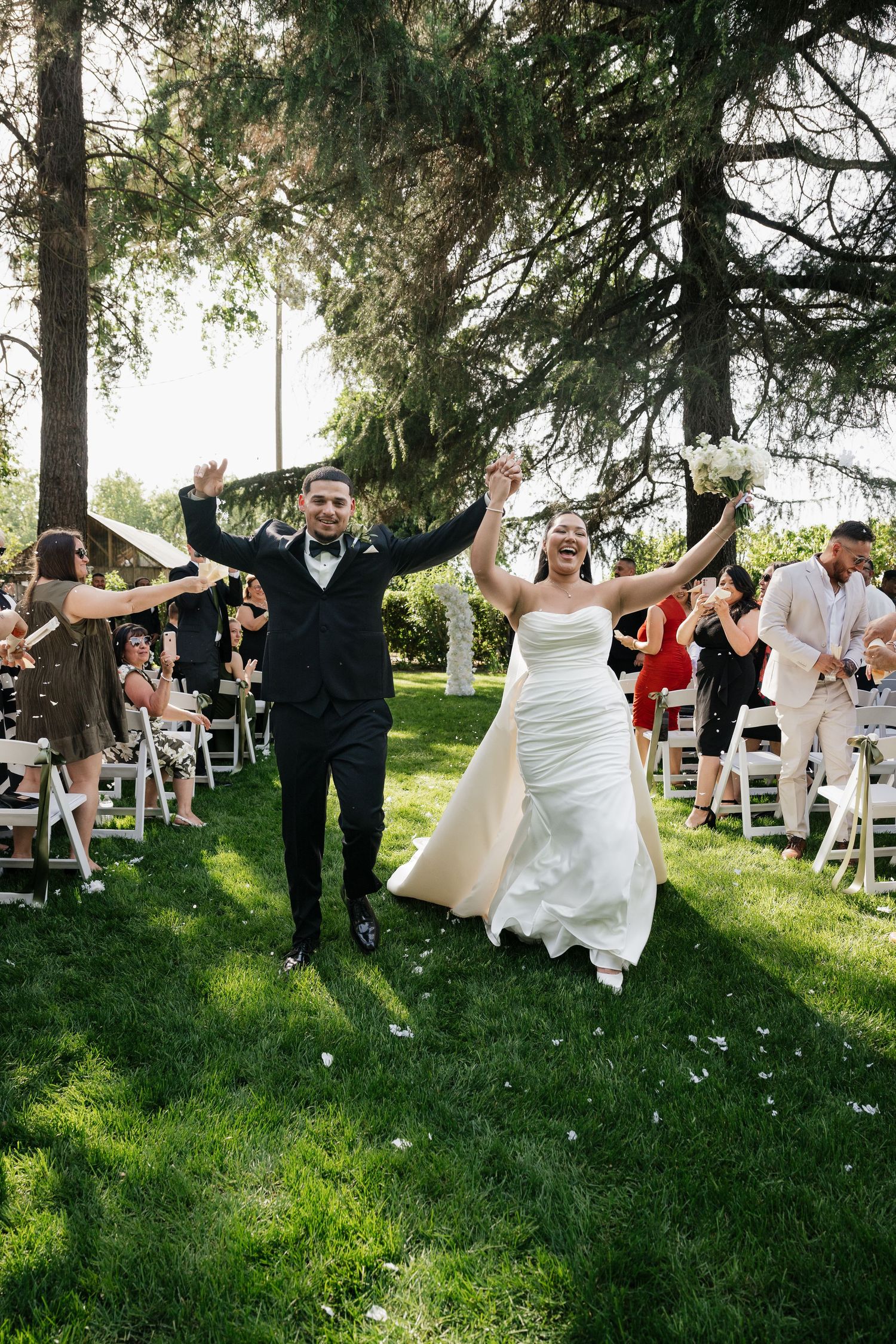 Joyful bride and groom walk down outdoor aisle, arms raised, surrounded by cheering guests on green lawn.