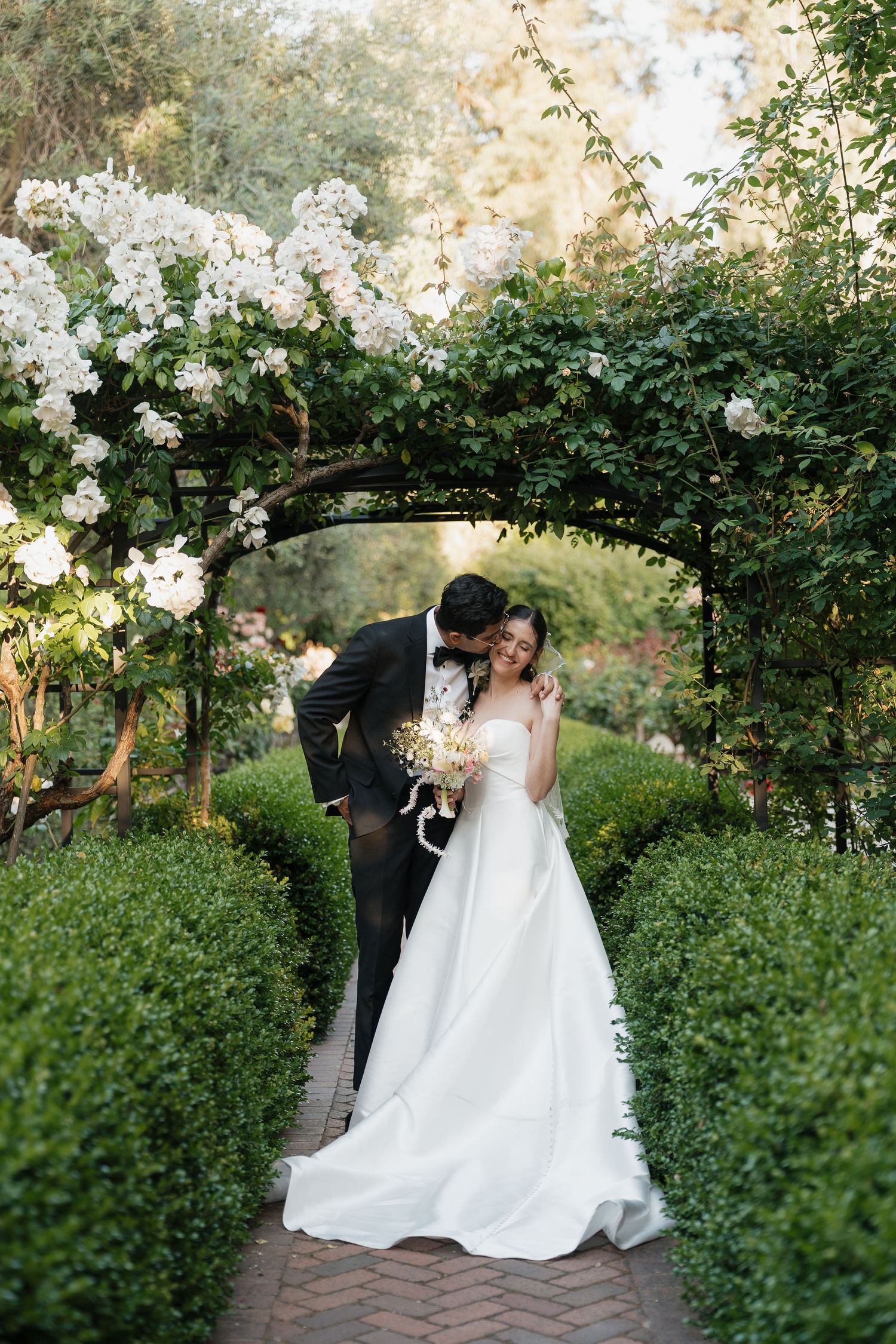 Bride and groom share a romantic kiss under a floral garden arch surrounded by lush greenery and white roses.