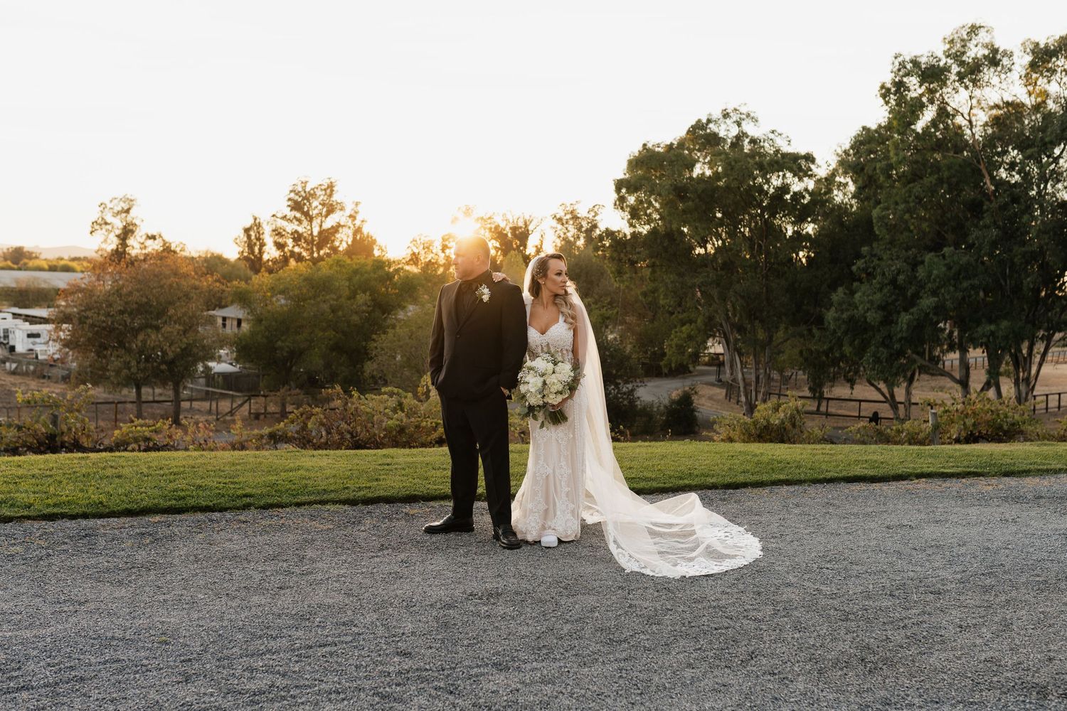 A wedding couple poses together on a pathway with dramatic sunset lighting and trees in the background.