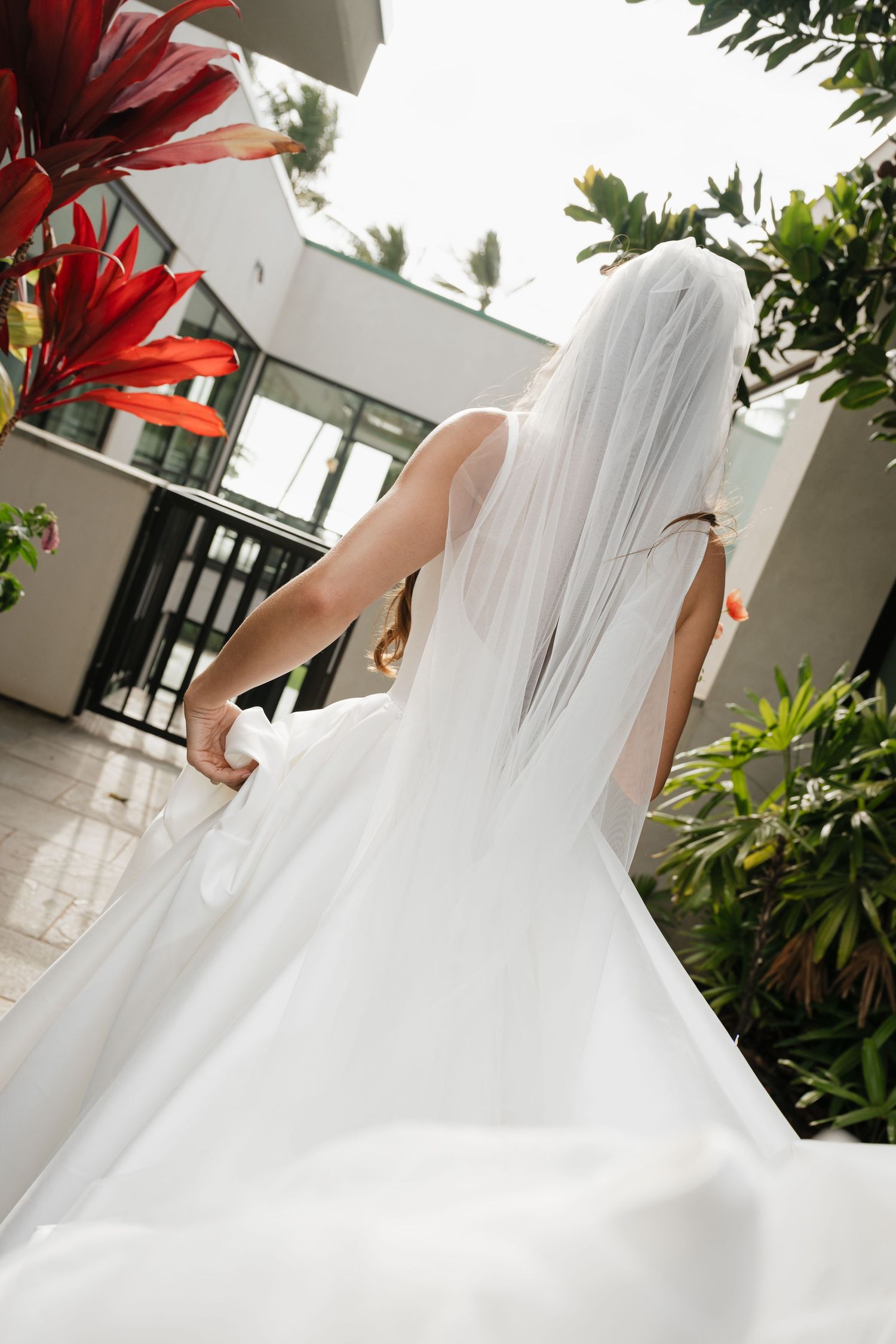 A bride in a flowing white wedding dress stands on a tropical balcony with red foliage visible in the background.