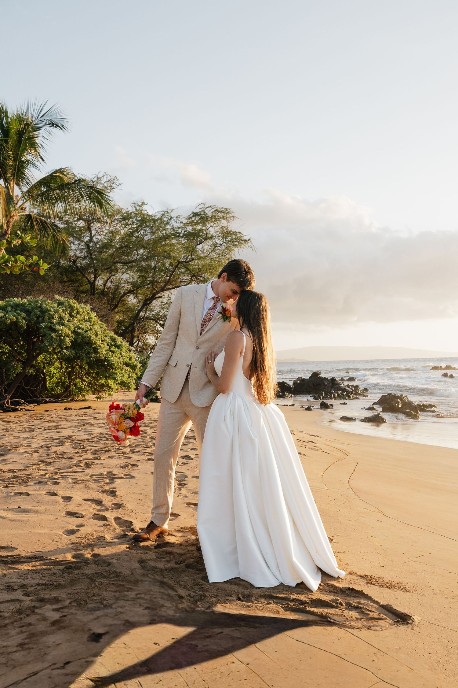 A couple in white attire shares a romantic moment on a scenic beach at sunset with palm trees in the background.