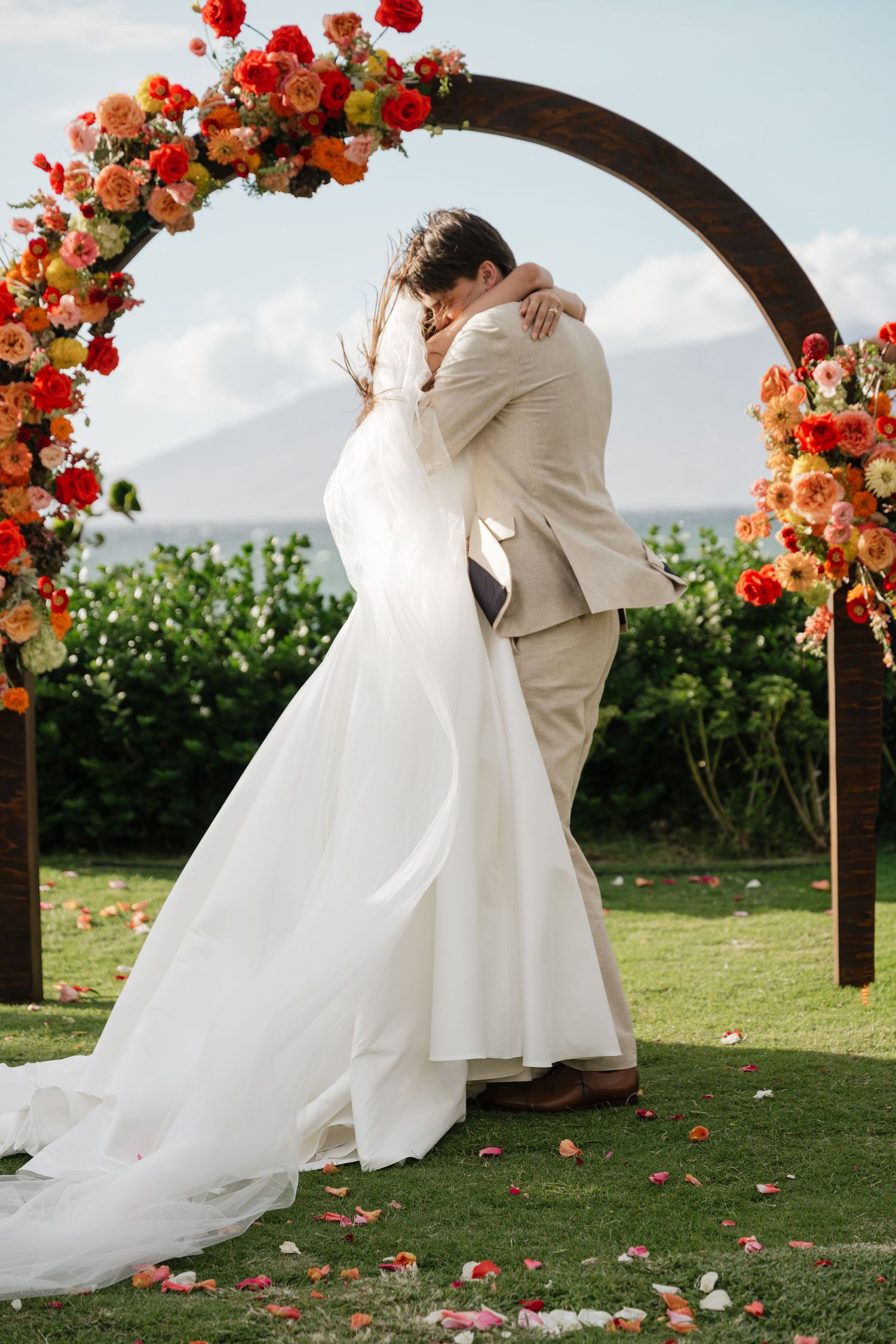 Wedding couple shares a romantic moment under a floral arch with red and orange roses against a scenic backdrop.