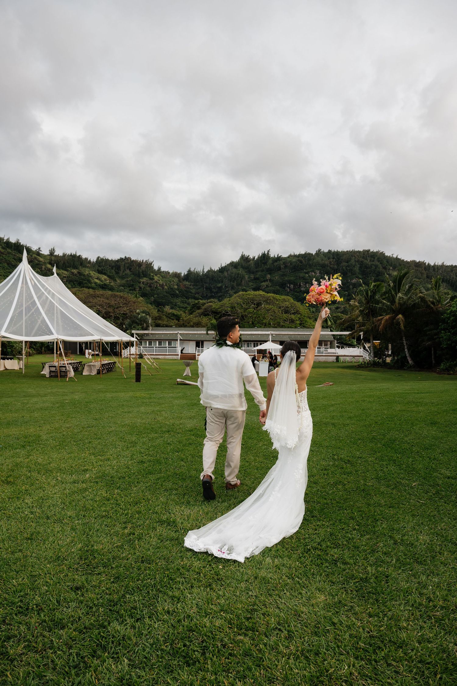 Bride and groom walking hand-in-hand toward white wedding tent on green lawn in Hawaii.