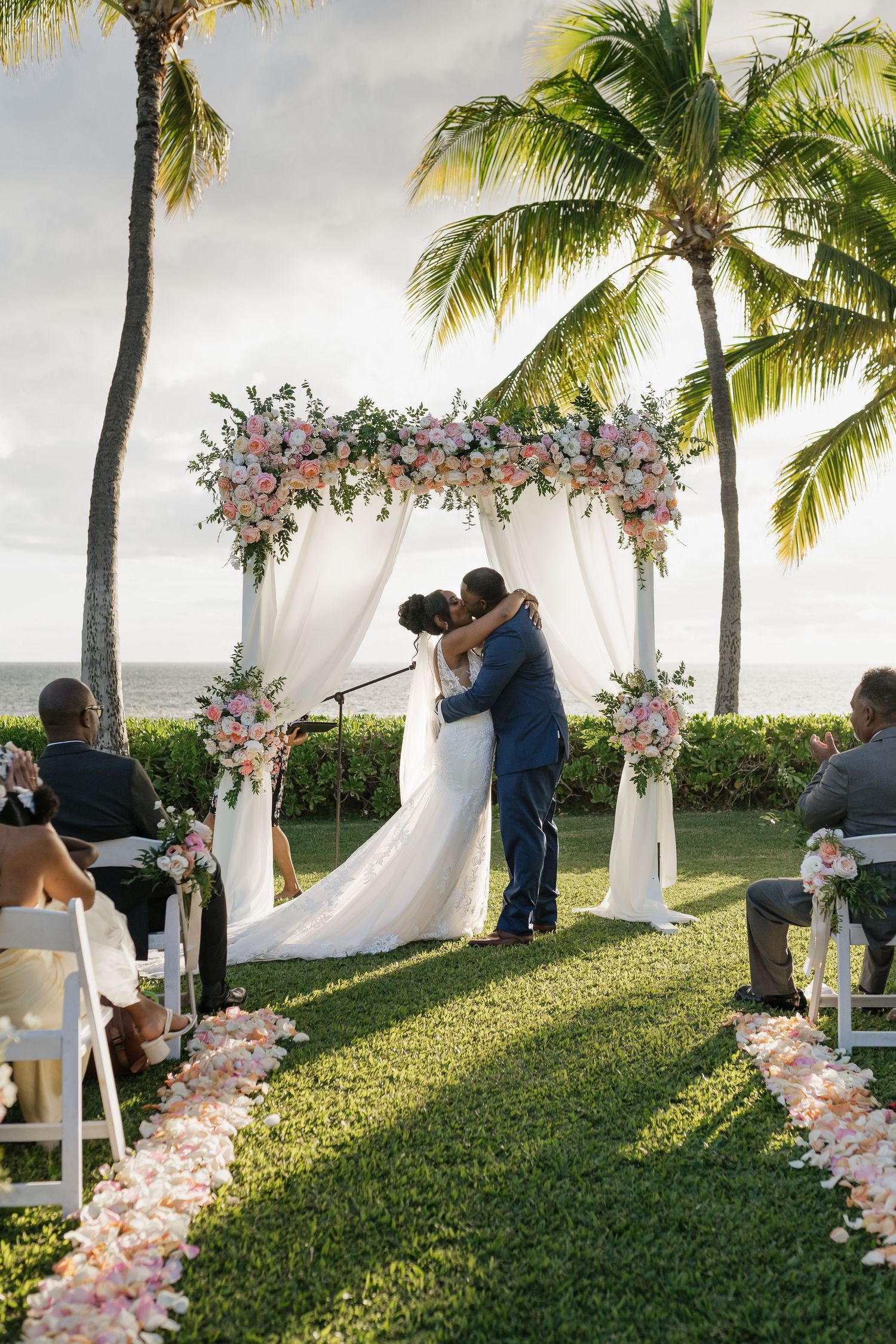 Outdoor wedding ceremony taking place under a floral arch with palm trees in the background.