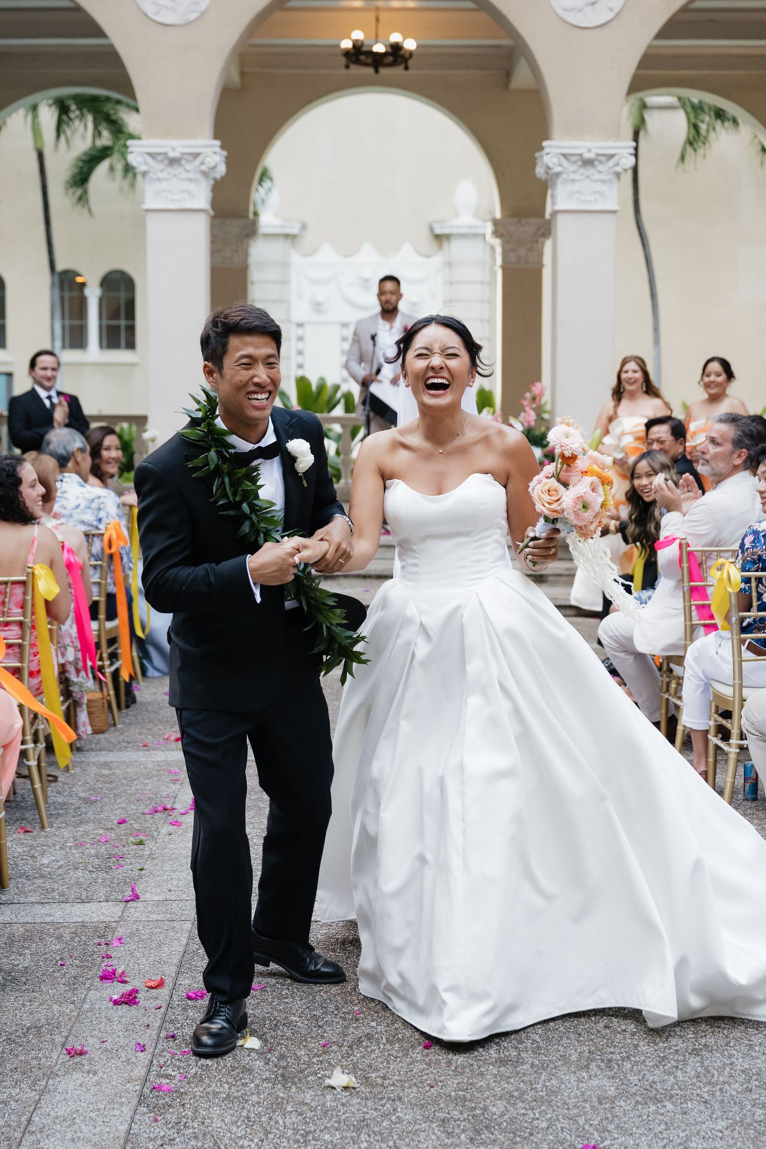Joyful newlywed couple walks down the aisle after wedding ceremony at elegant outdoor venue with guests cheering.