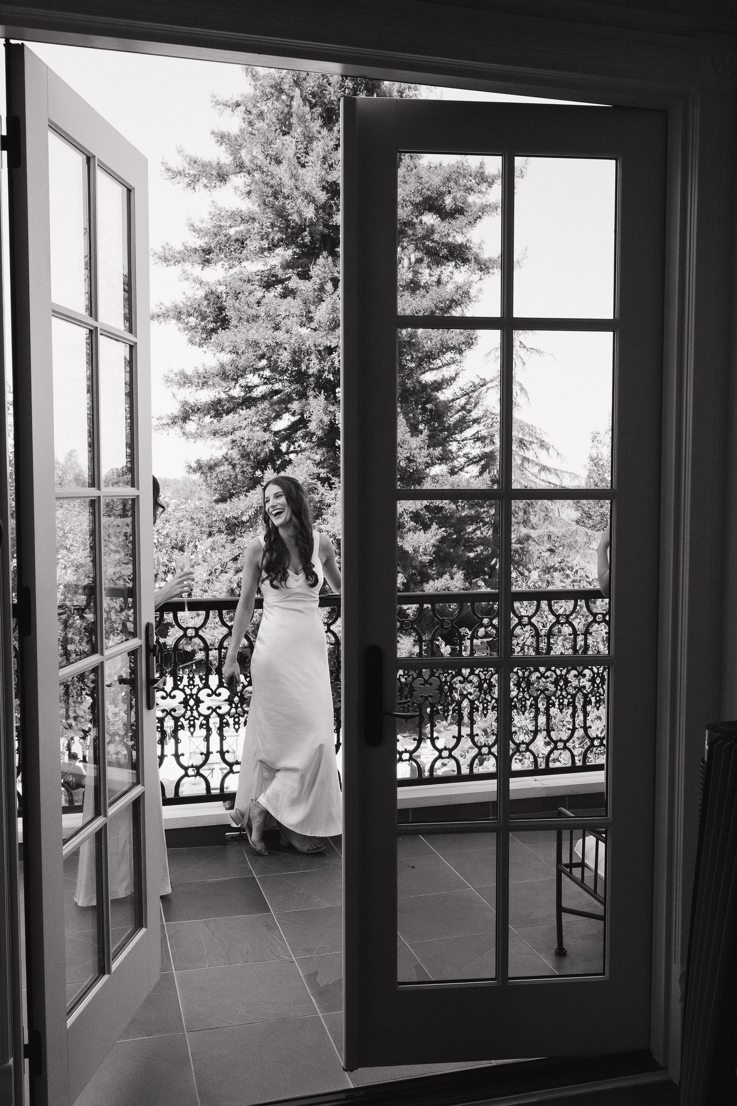 Black and white photograph of a wedding dress silhouetted against large windows with ornate balcony railings.