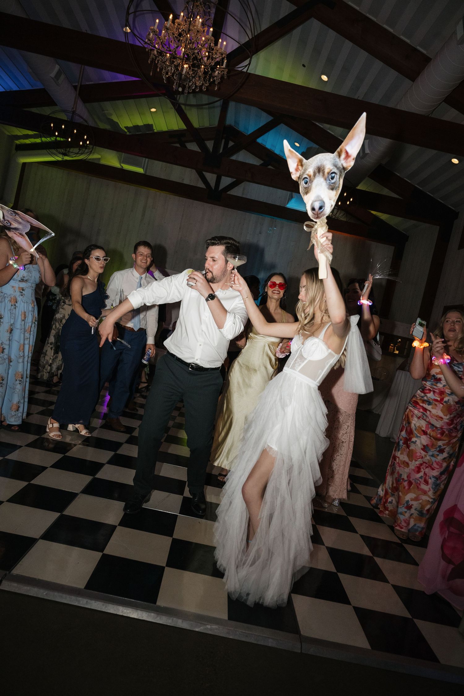 Wedding reception dance floor with guests celebrating on black and white checkered tiles under wooden beams.