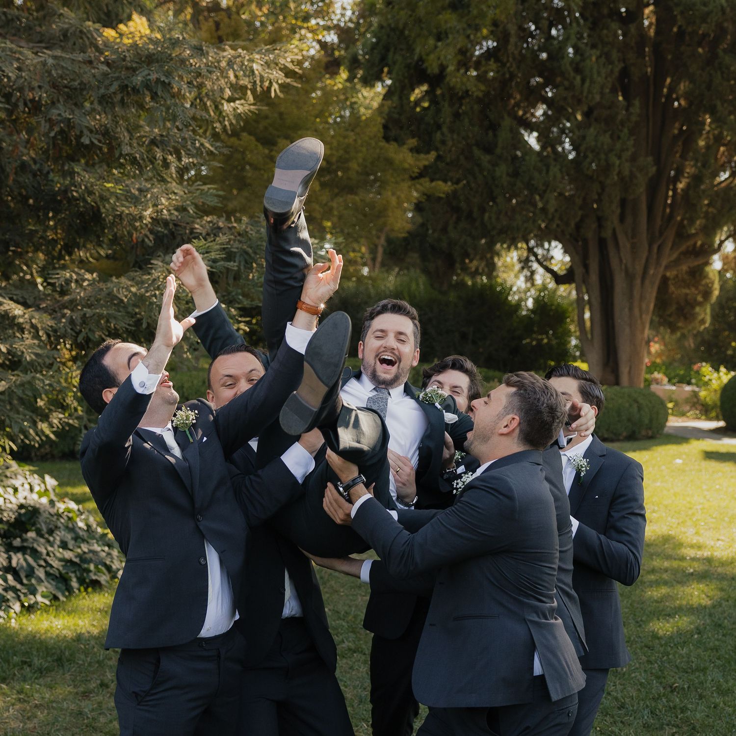 Groomsmen in black suits playfully pose for a sequence of candid celebration shots outdoors in a garden setting.