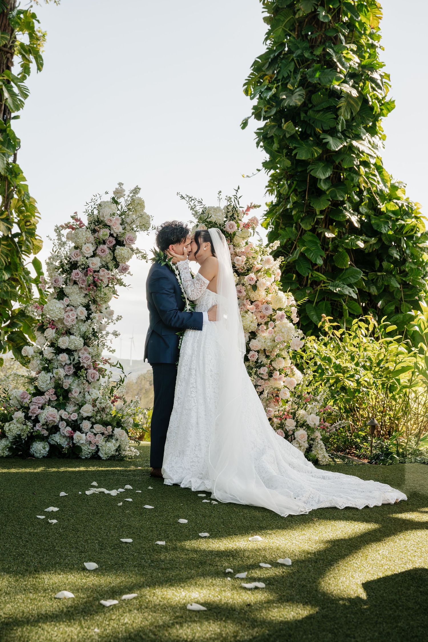 Bride and groom share first kiss under floral arch with white and pink blooms at outdoor wedding ceremony.