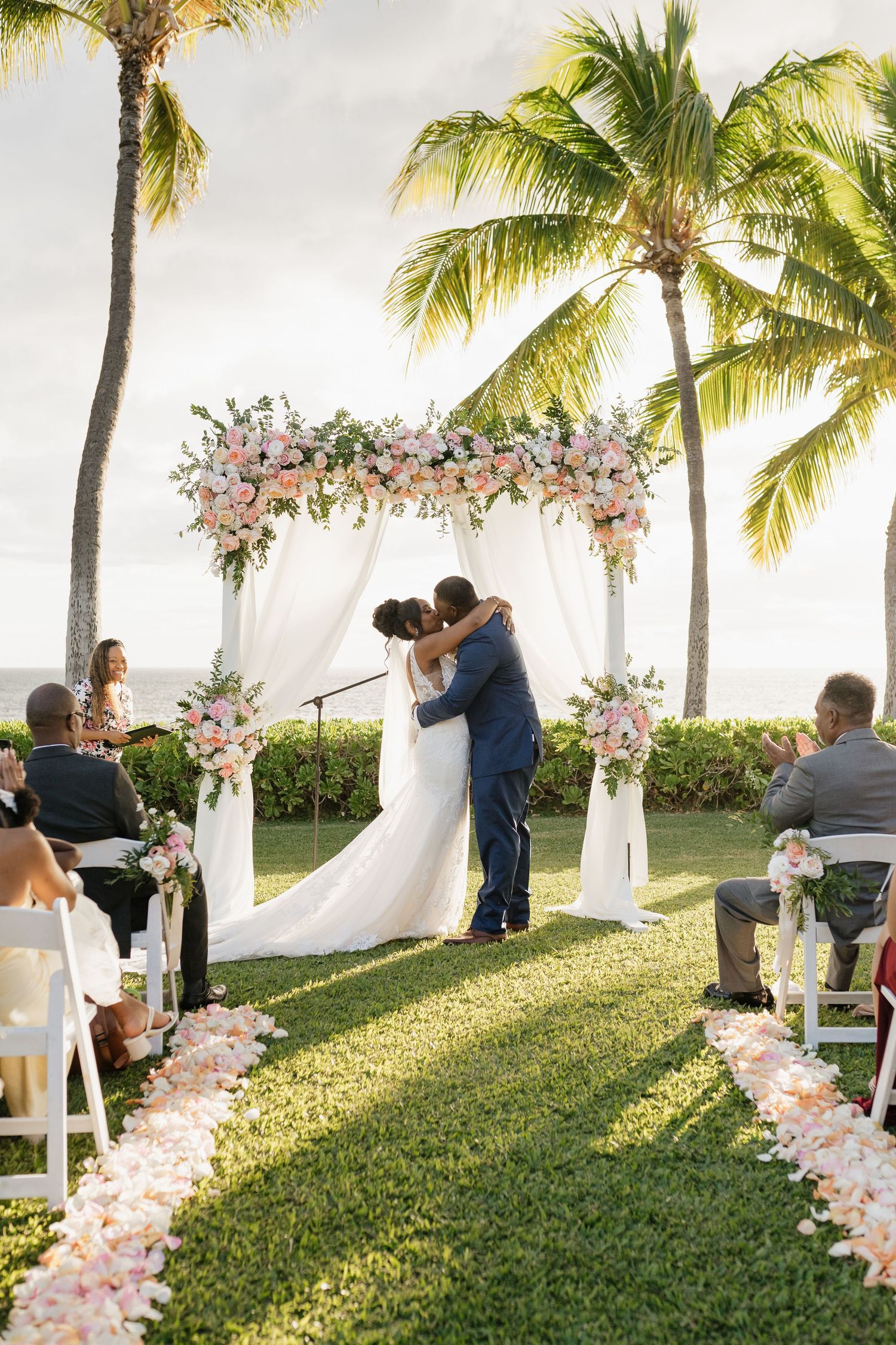 Wedding ceremony on tropical lawn with palm trees and white floral arch.