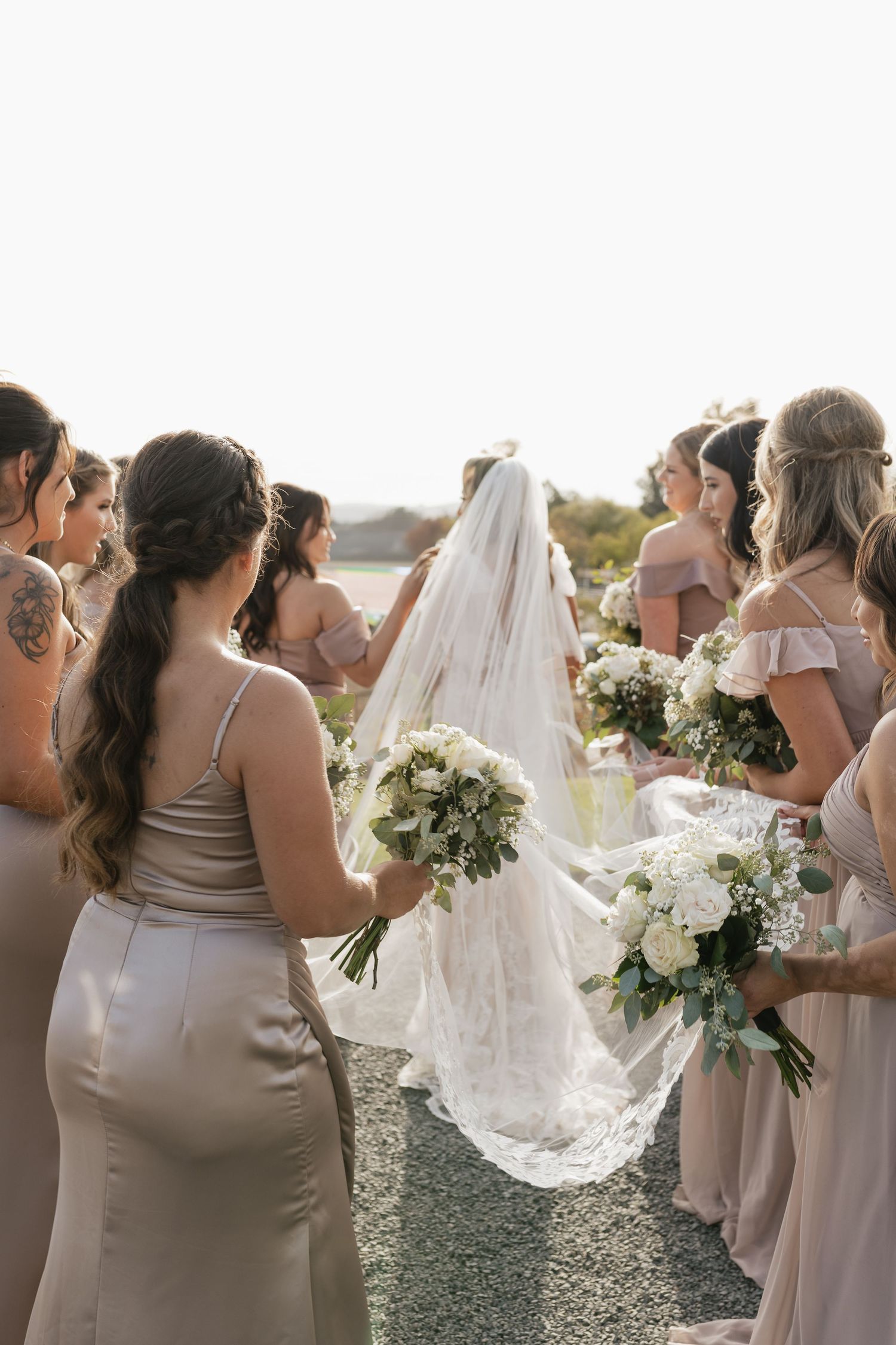 Wedding party in neutral-toned dresses gathered outdoors holding white bouquets.
