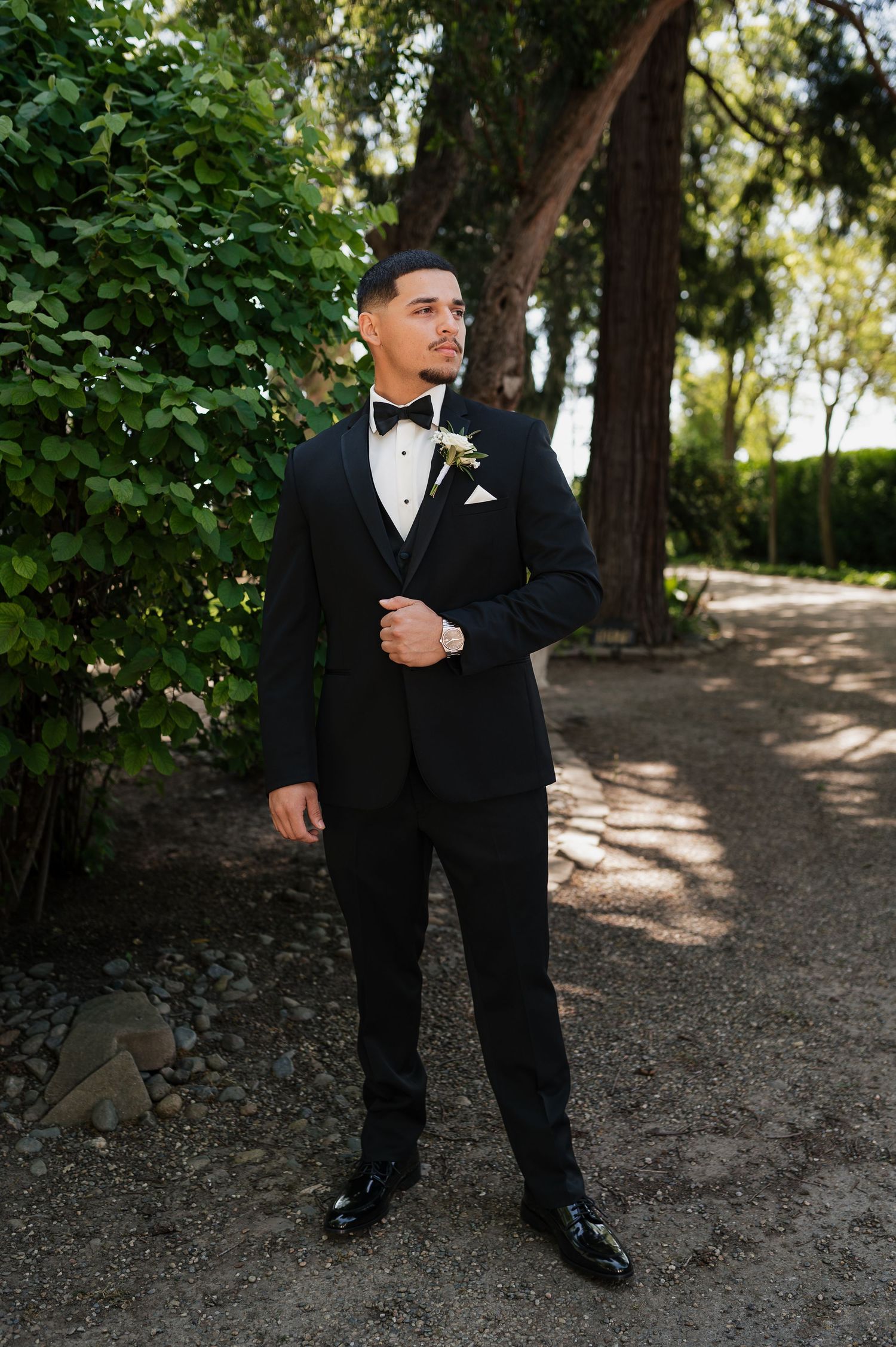Groom in black tuxedo with boutonniere stands on garden path surrounded by lush greenery on wedding day.