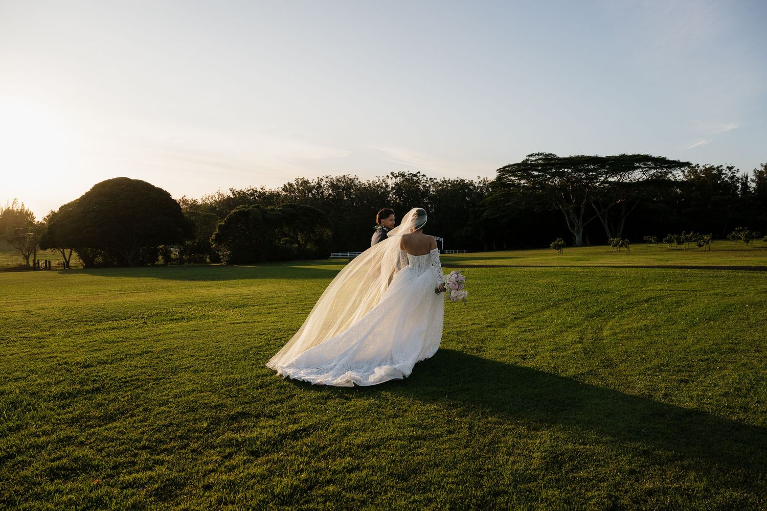 Bride in flowing white wedding gown and veil walks across lush green lawn at golden hour sunset.